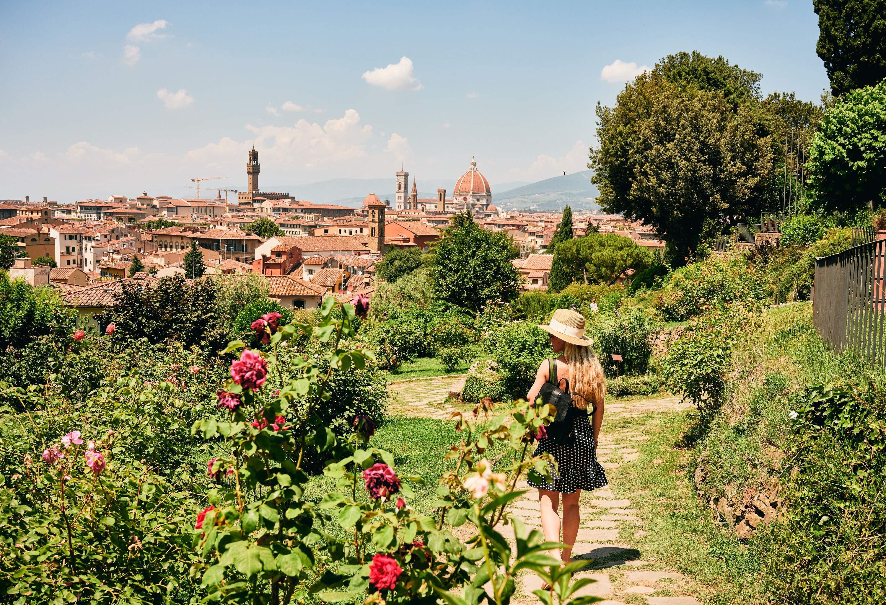 A curly blonde lady walks in a cobbled path of grass surrounded by fresh flowers and plants with a cityscape view.