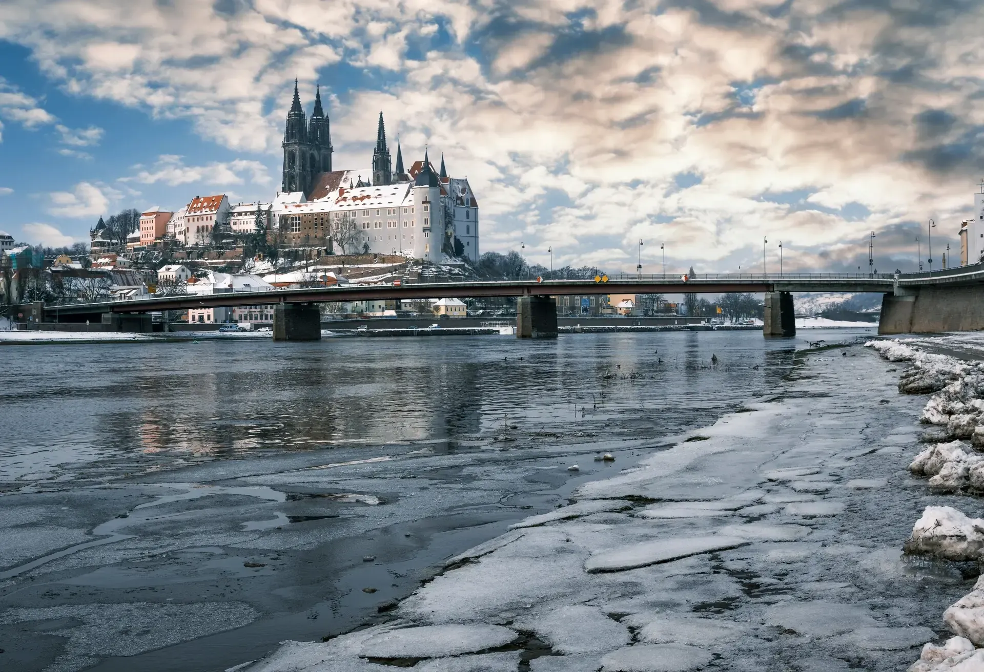A wintery landscape featuring a historic European town with a large cathedral and castle atop a hill. A bridge spans a partially frozen river in the foreground, with patches of ice and snow visible along the riverbank. 