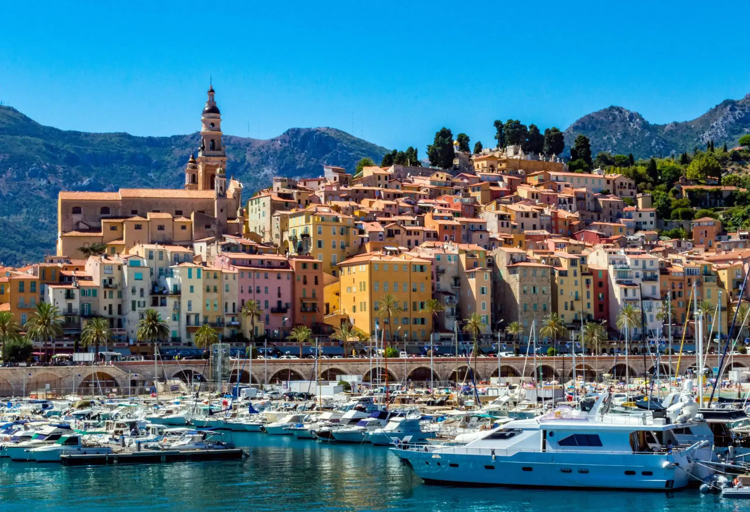 A packed harbour with colourful hillside houses along a roadway on the coast.