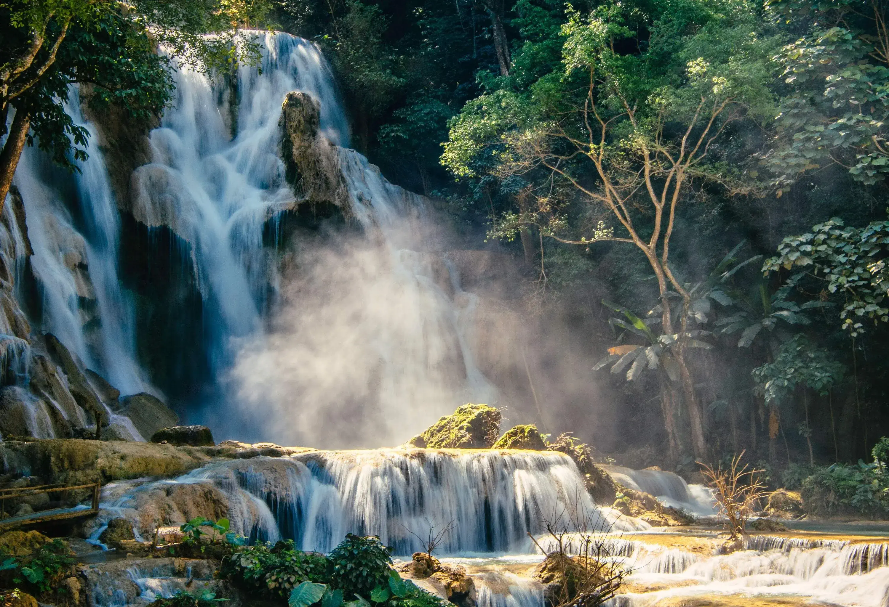 A beautiful waterfall cascades into the tiered rocky streams surrounded by lush trees.