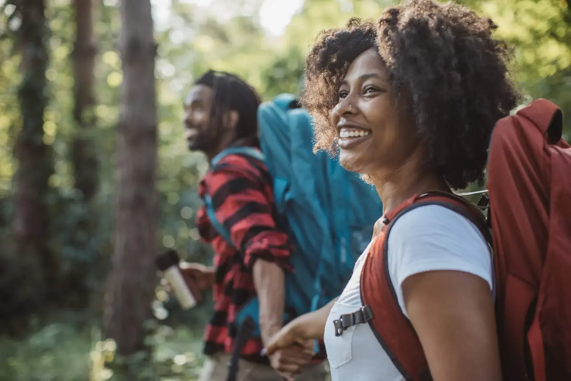 Smiling couple in a woodland holds hands and carries large hiking bags.