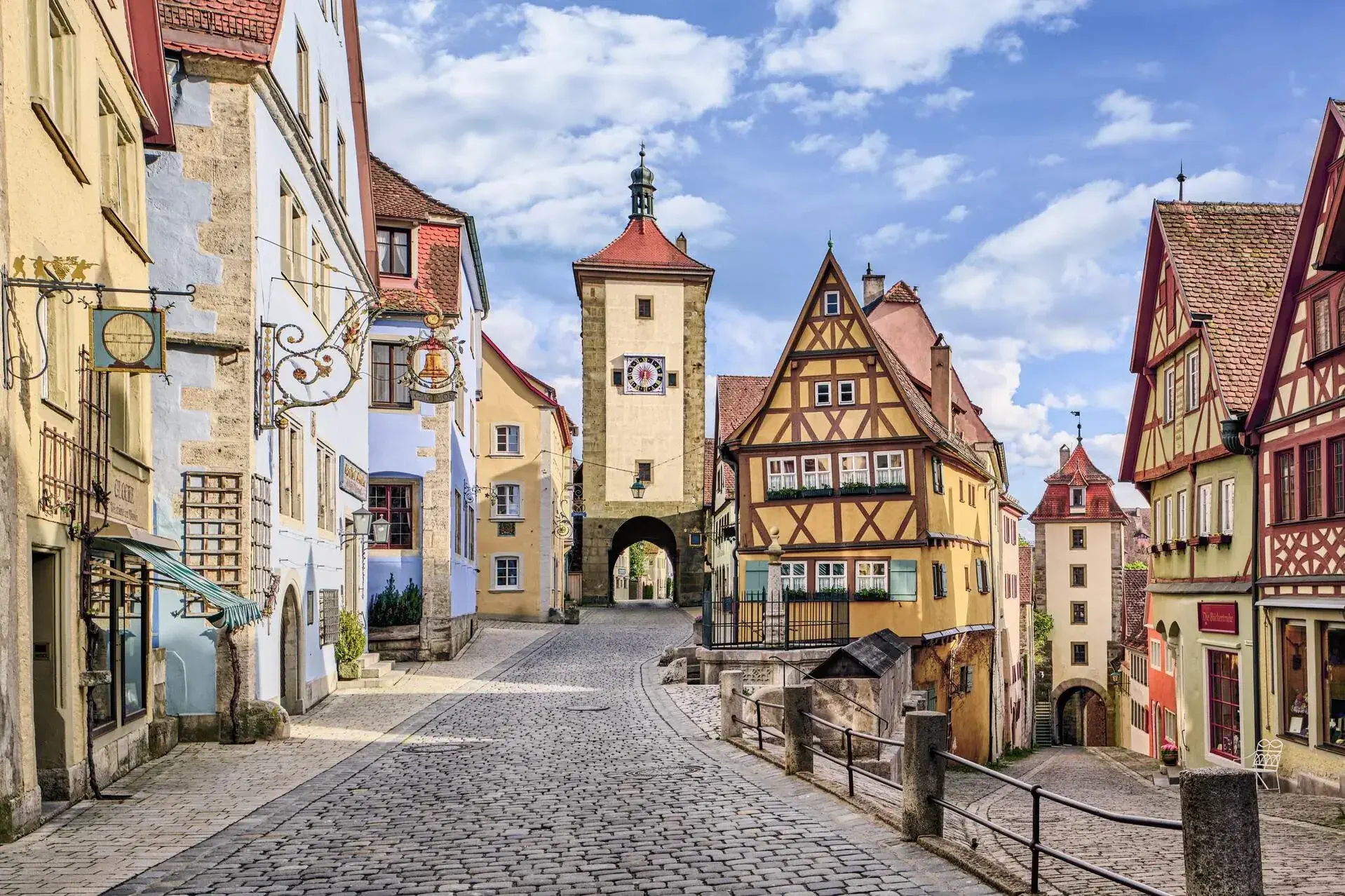 A brick road with steel railings and several half-timbered homes on either side leads to a gateway. 