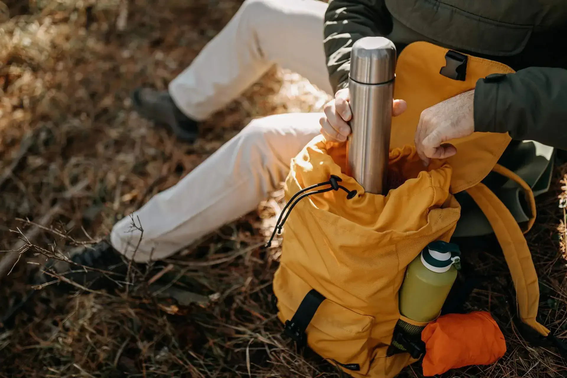 A man resting on the ground brings out a silver vacuum-insulated bottle from his yellow backpack.