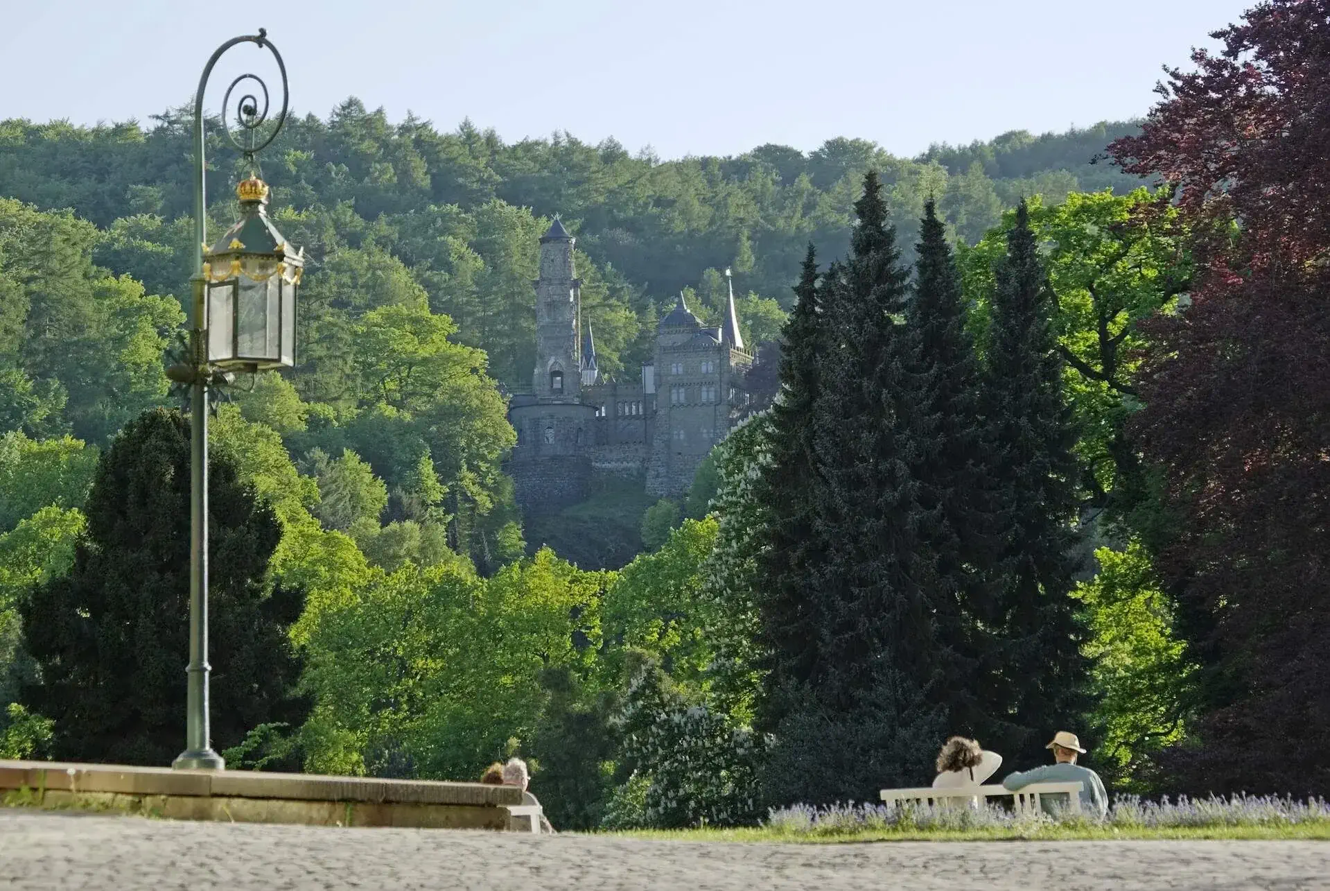 Four people sitting on benches in a green park and enjoying the view of a castle between the trees