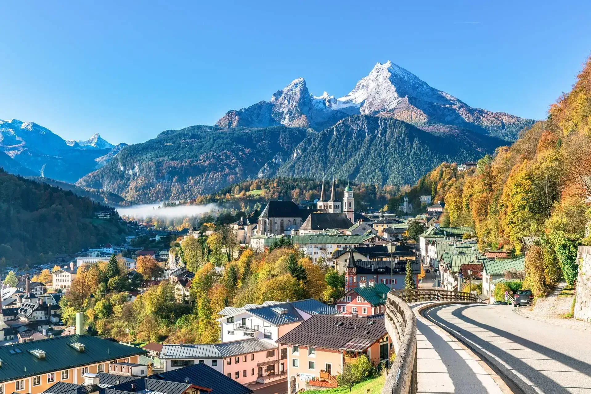 A hillside road overlooking a village amongst autumnal trees at the base of snow-capped mountains.