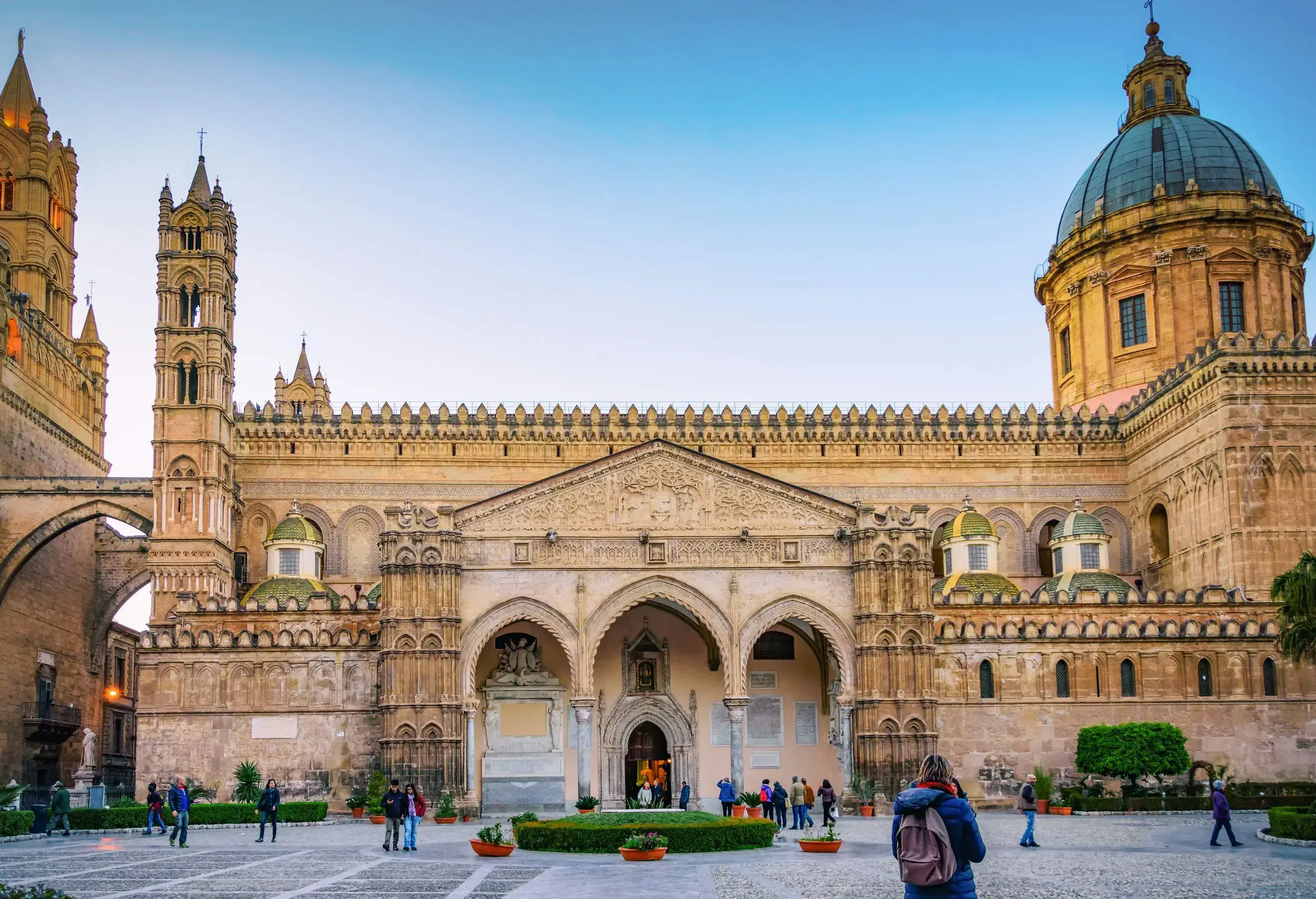 Palermo Cathedral is a church with a dome roof and intricately carved façade with a bell tower and portico in front of a busy plaza.