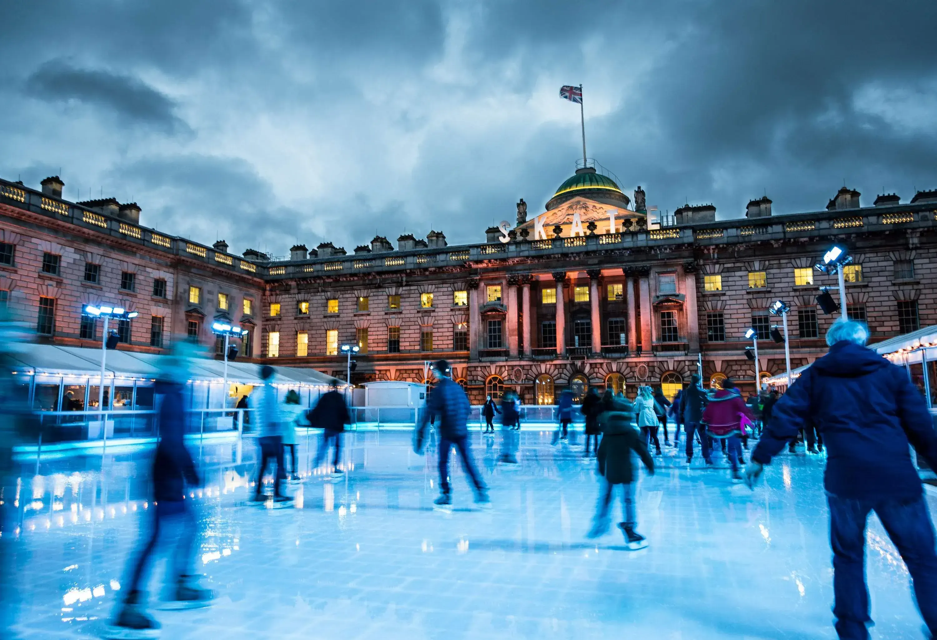 Blurred motion of crowds of people ice skating at Somerset House, a publically owned building in central London. The image features warm evening light, and an ominous, moody sky. The people appear as unrecognisable blurs due to the long exposure used. Somerset House is a large Neoclassical building situated on the south side of the Strand in central London, overlooking the River Thames, just east of Waterloo Bridge.