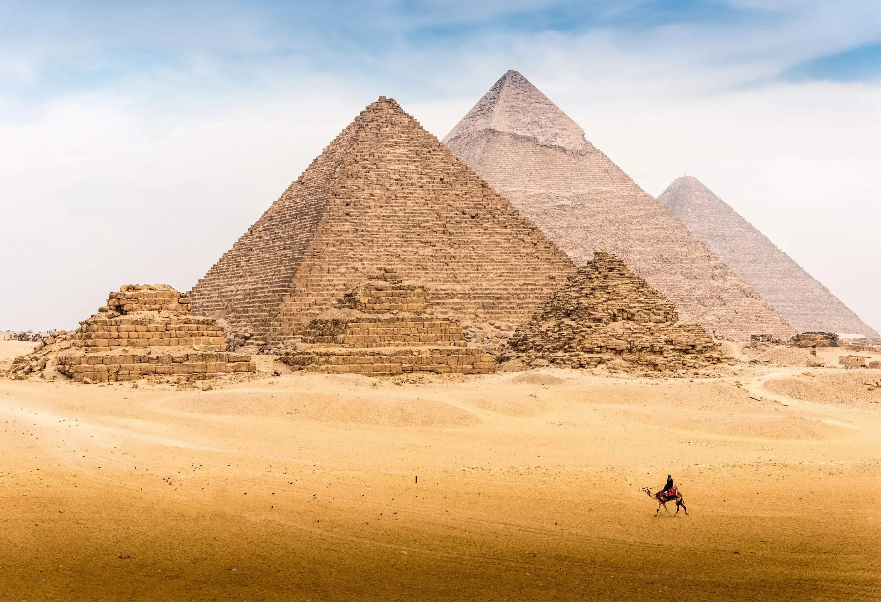 A man riding a camel passes through the iconic ancient pyramids protruding into the sky.