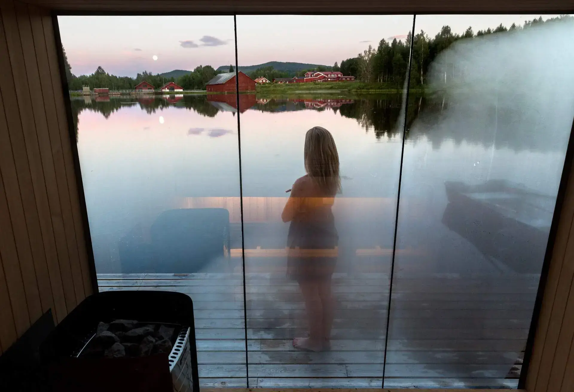 Woman standing outside a floating sauna overlooking a lake, photographed from inside at dusk