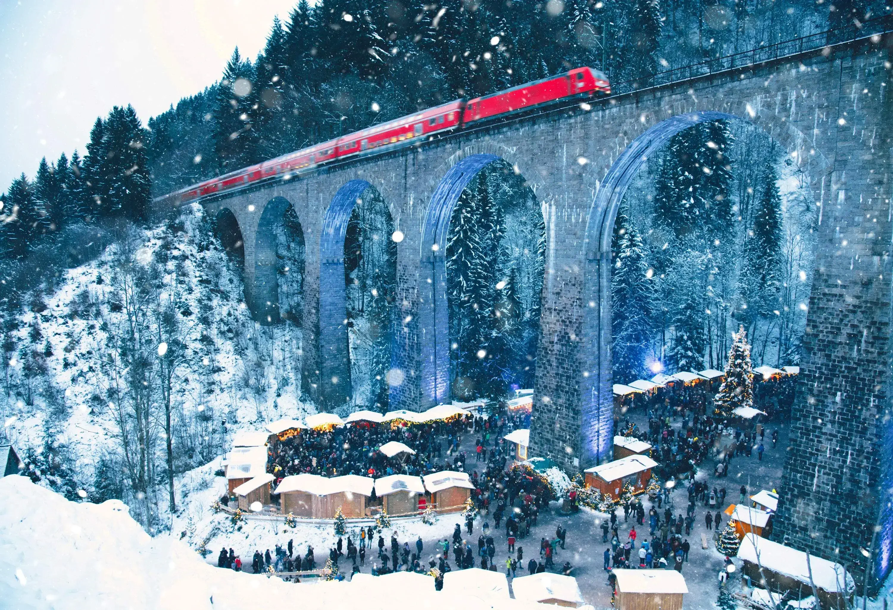A crowded Christmas market with stalls beneath a viaduct with a passing train during snowfall.