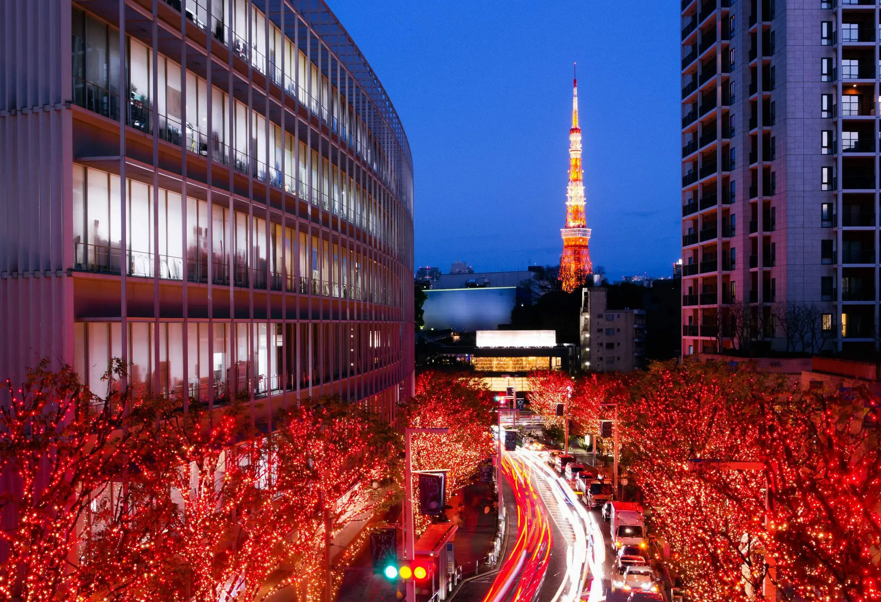 Cars driving through a busy street surrounded by trees wrapped in red lights and glass-cladded buildings with views of the glowing Tokyo Tower.