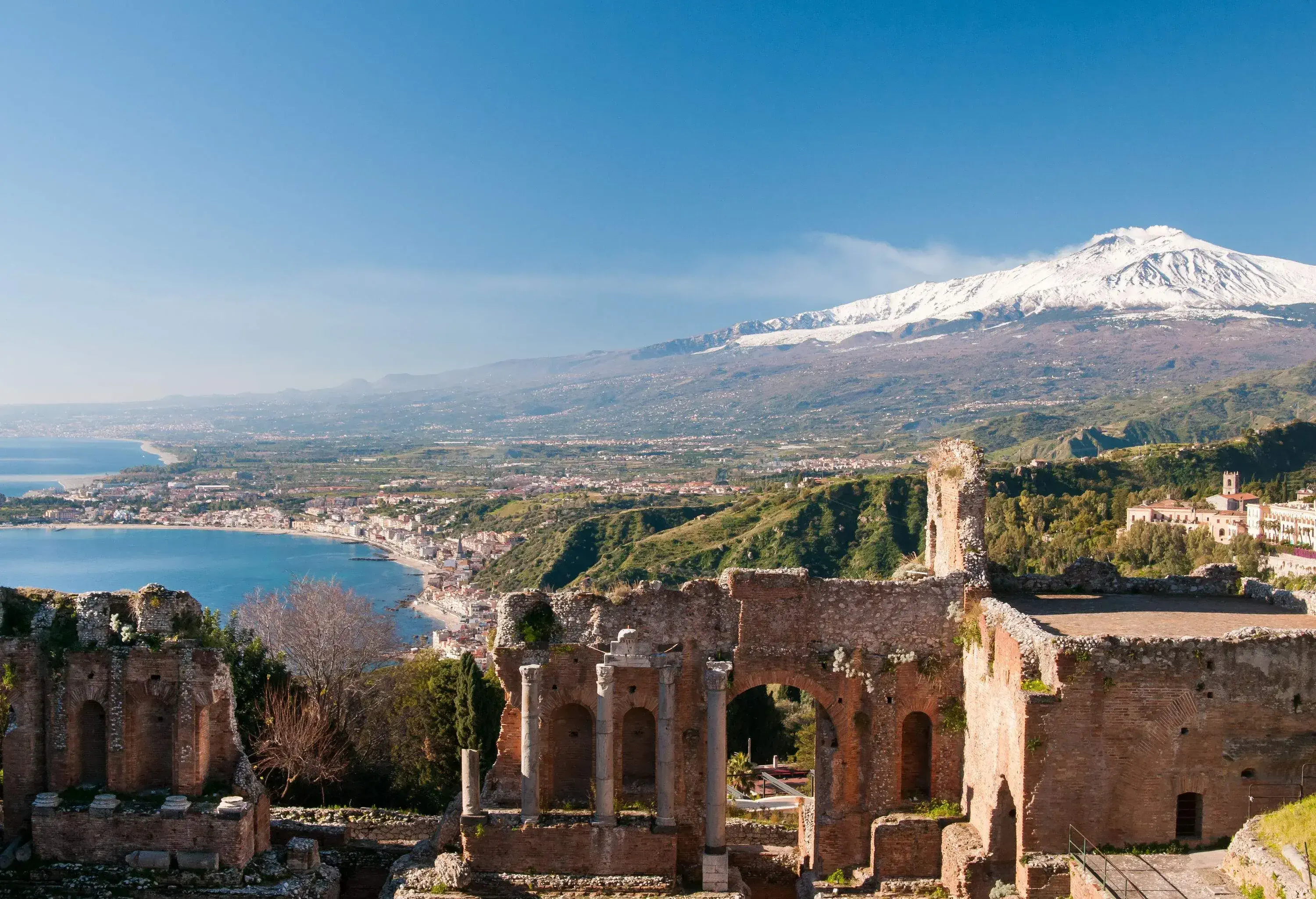 Ruins of an ancient Greek theatre on a hilltop with a scenic view of the snow-covered Mount Etna and the sea.
