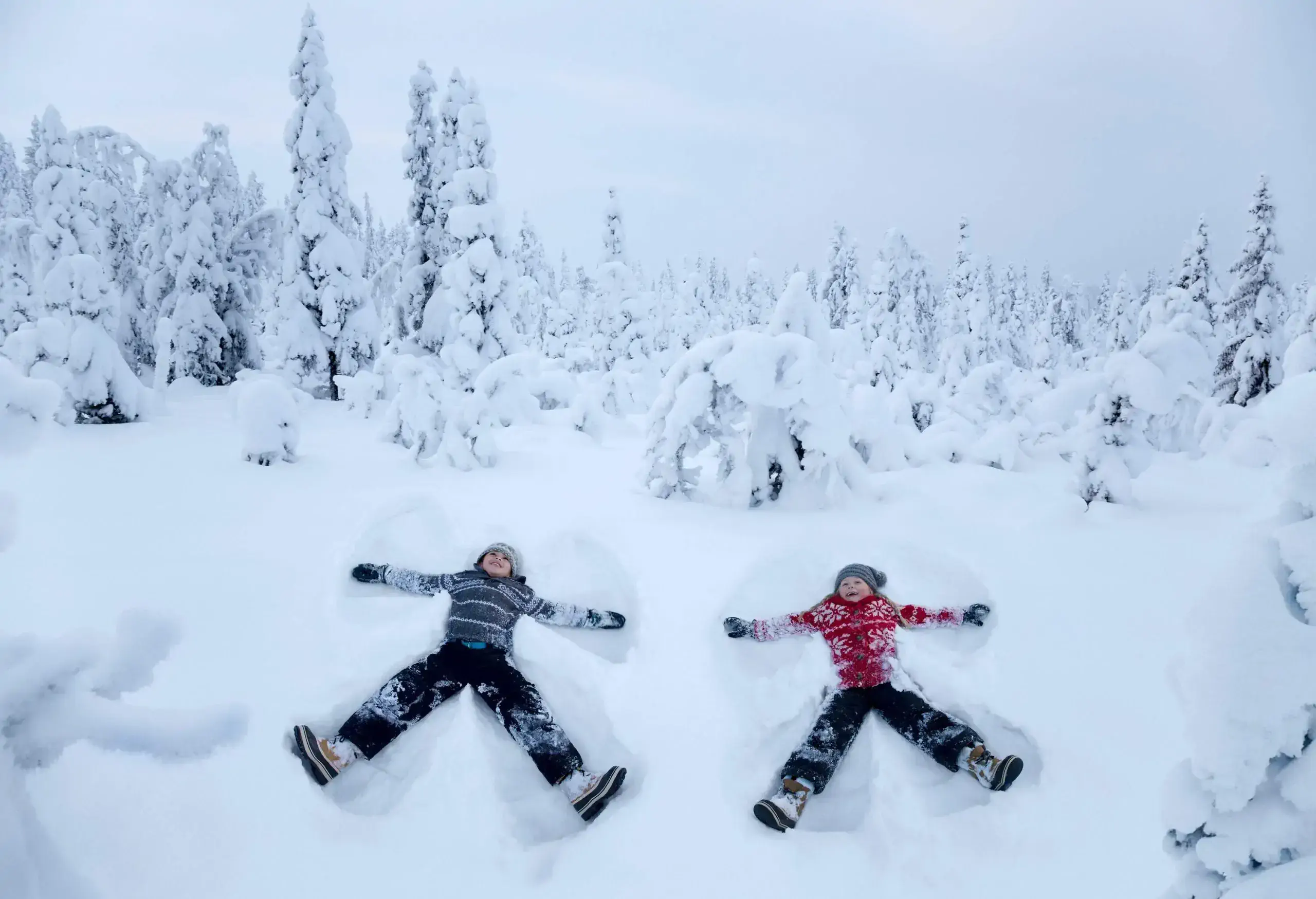 Two kids making snow angels in the snow, with heavily frosted trees behind them.