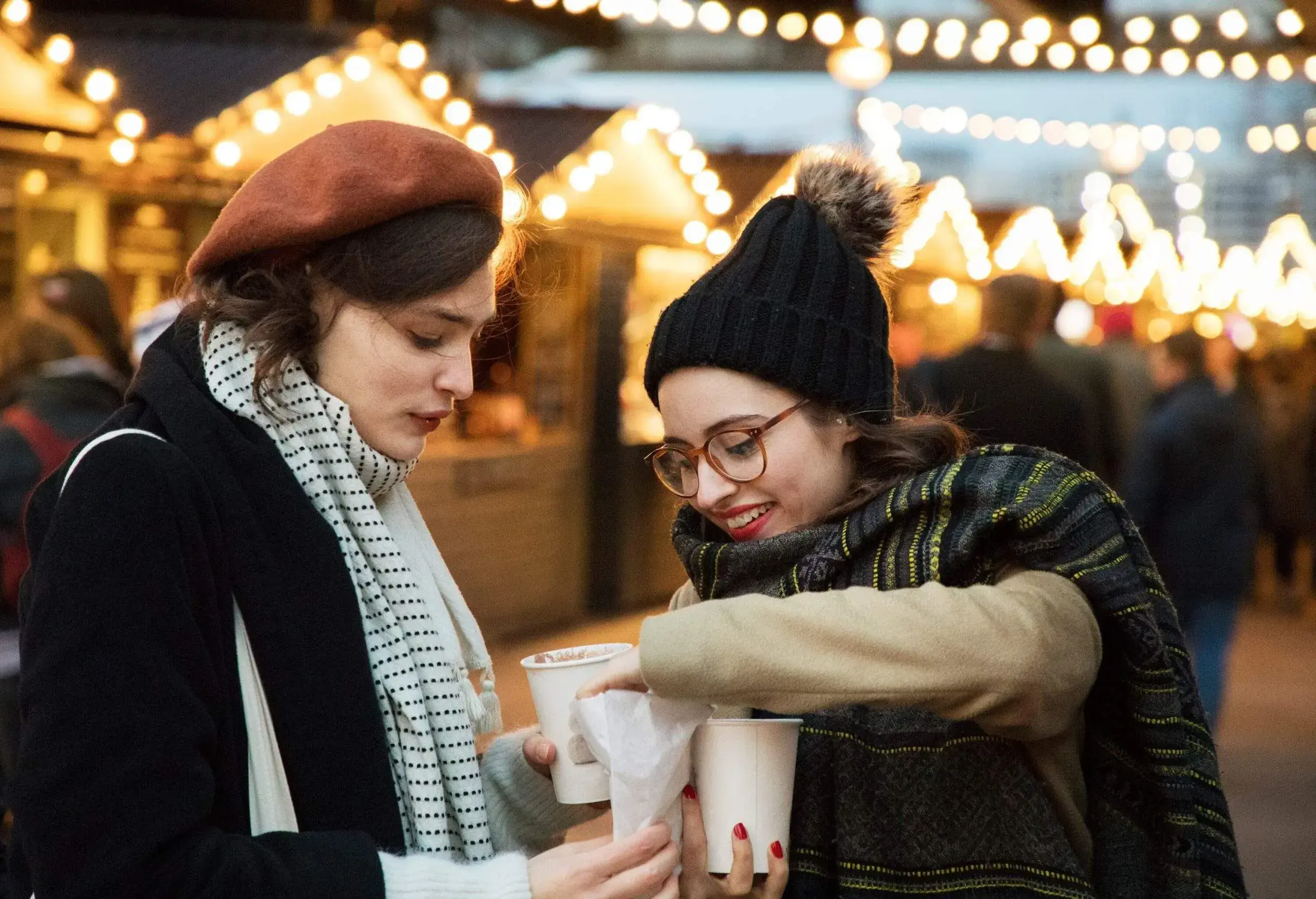 two women in winter attire enjoy hot beverages at an outdoor market with festive string lights in the background.