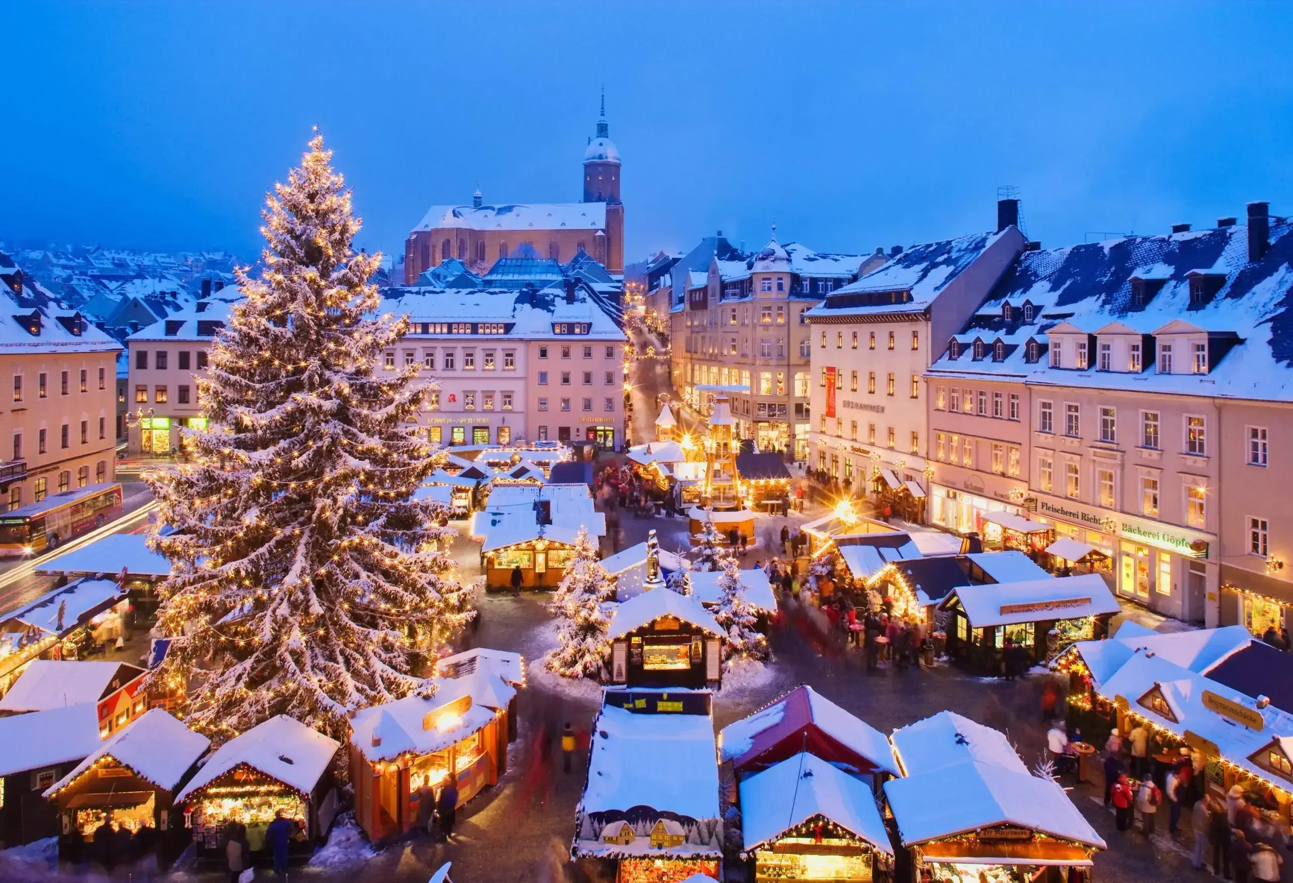 A busy Christmas market with a large Christmas tree in the middle surrounded by city structures at night.