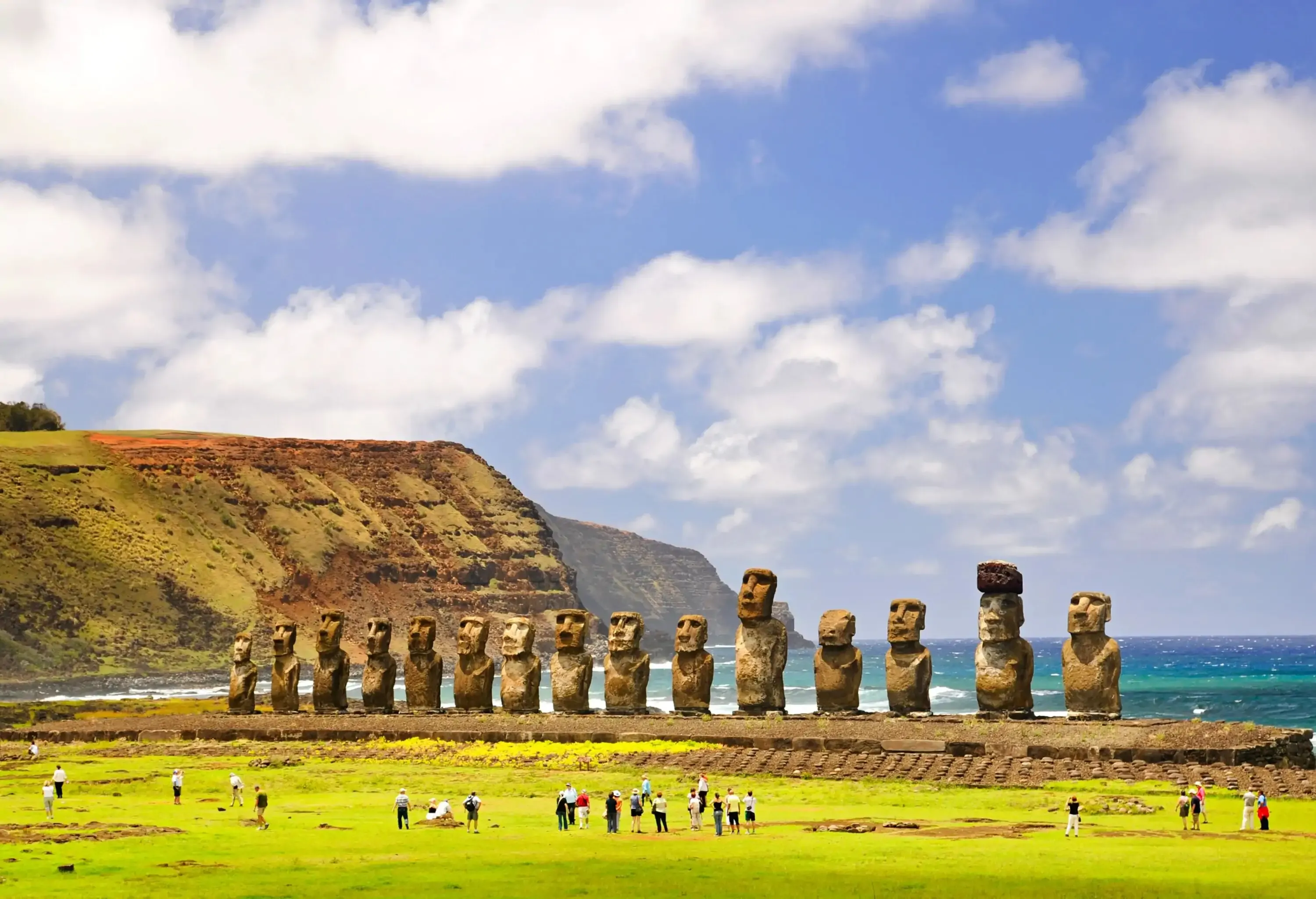 The iconic 15 Moai statues, large stone monoliths carved by the Rapa Nui people, stand tall along the coastline, with visitors exploring the nearby turf and a dramatic coastal cliff serving as the backdrop.