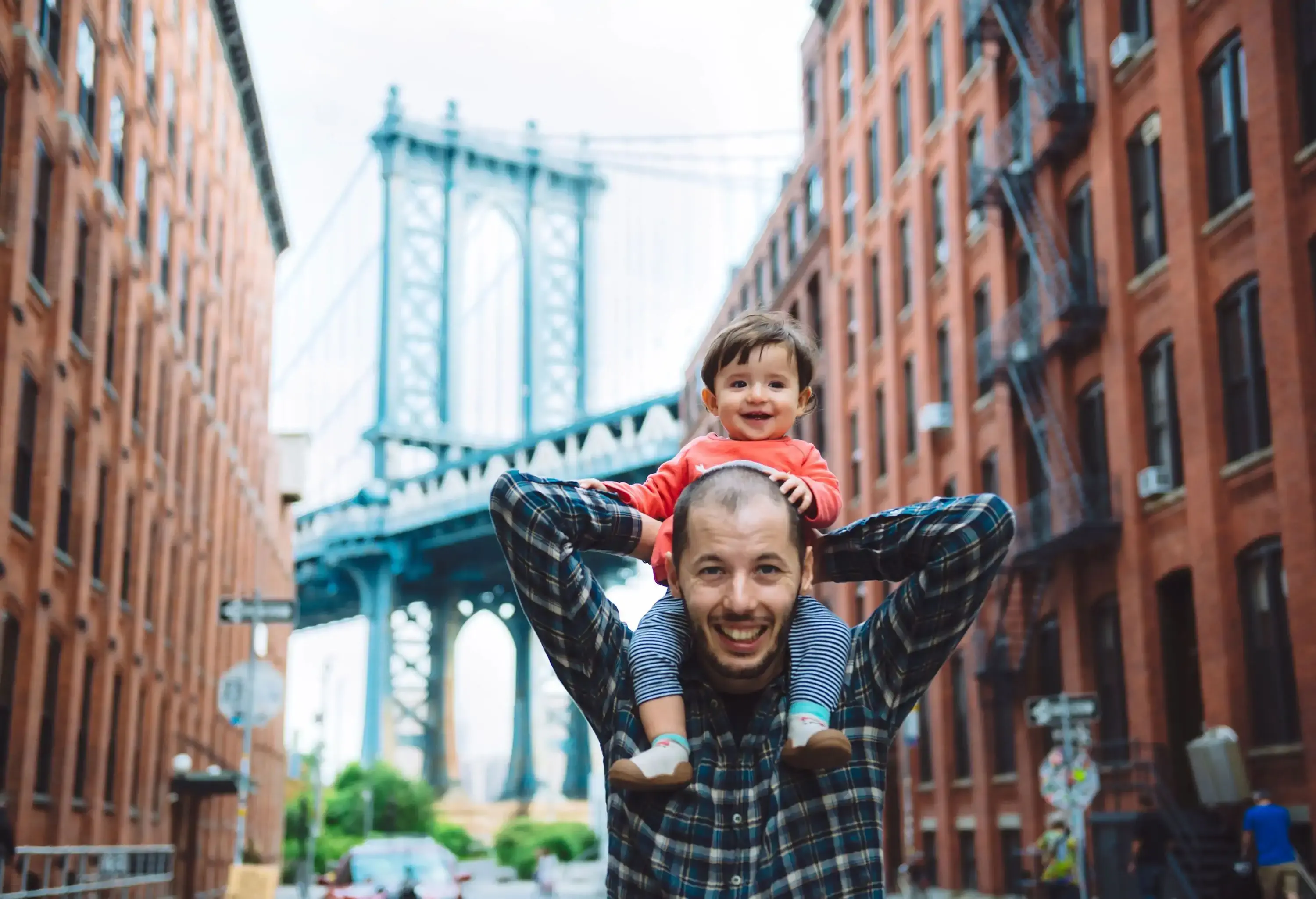 A man with a little boy propped over his shoulders standing in the street. with the Manhattan Bridge in the backdrop.