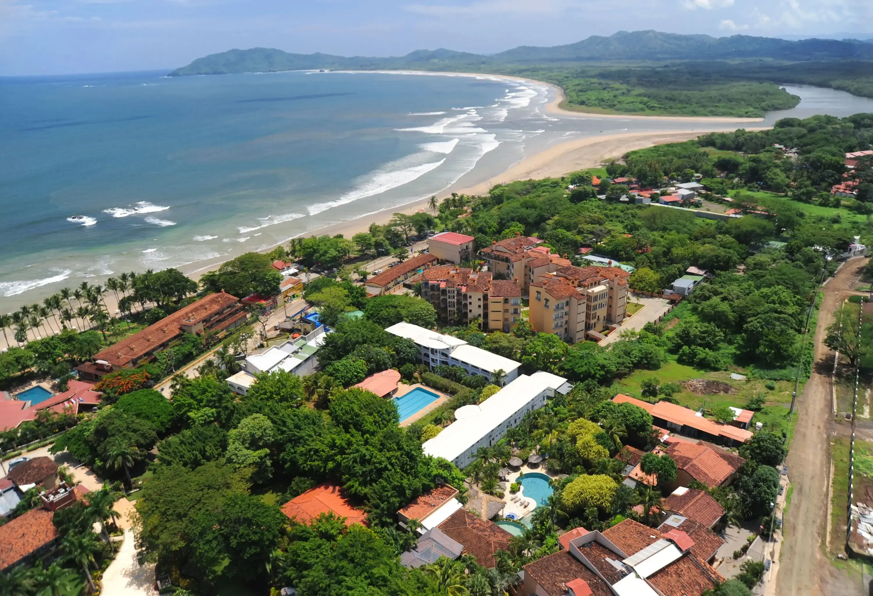 A sandy beach leads to a coastal town blanketed in trees and buildings, with a river mouth meandering into the scene from the background, merging land and sea in a captivating panorama.