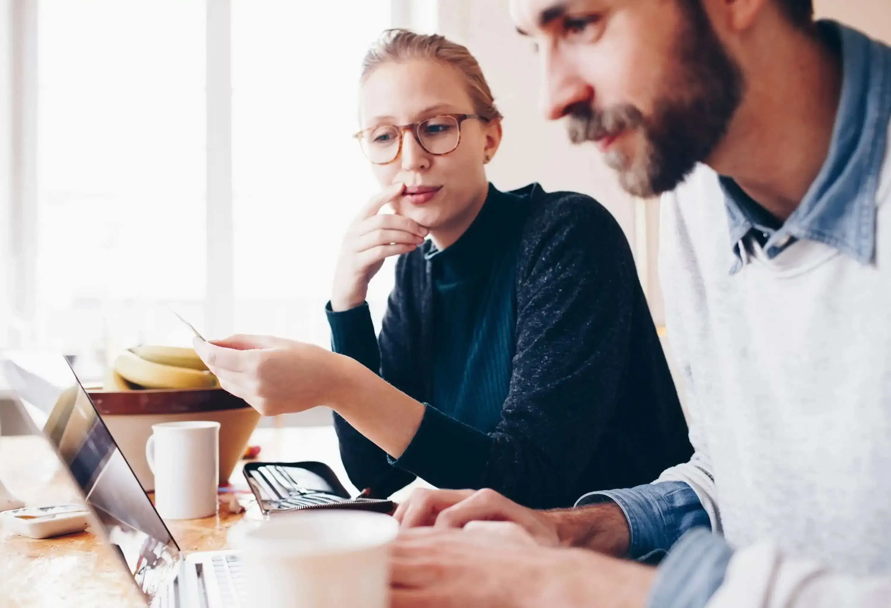 A couple talks at a table while the man works on his computer. 