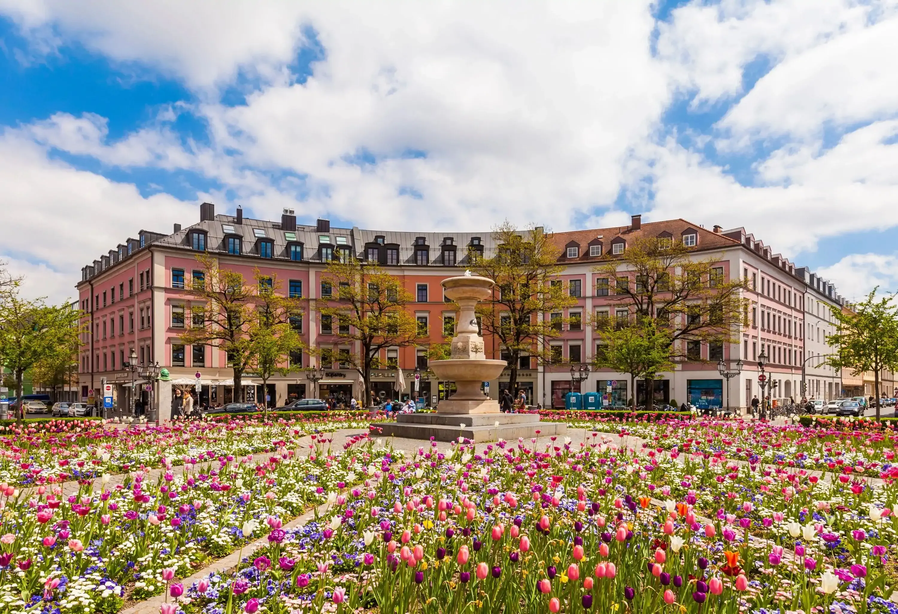 A central square with a fountain in the middle of colourful, lush flower beds surrounded by buildings.