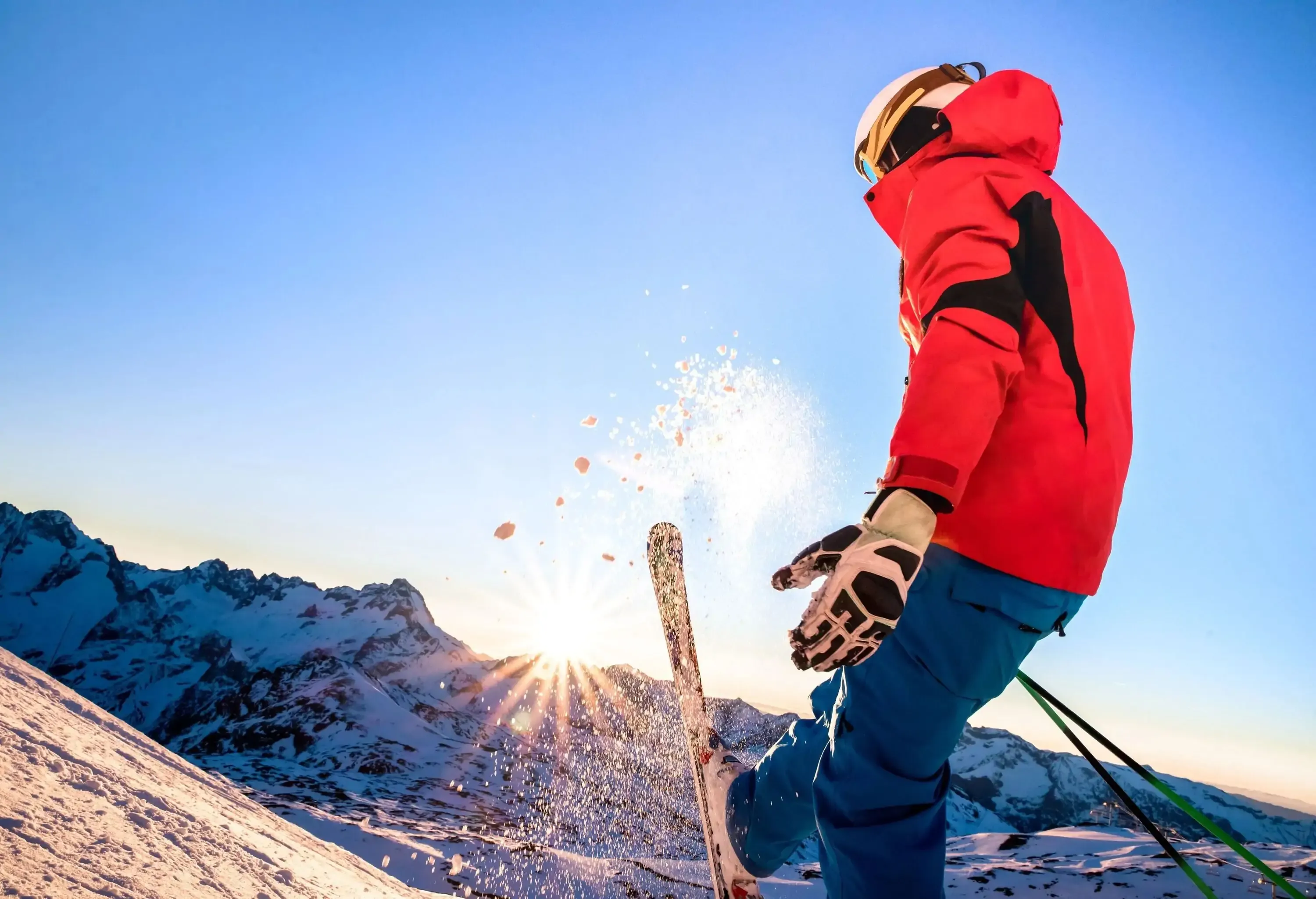 A skier in a red jacket slowing down on a slope as the light shines through the snowy mountain range.
