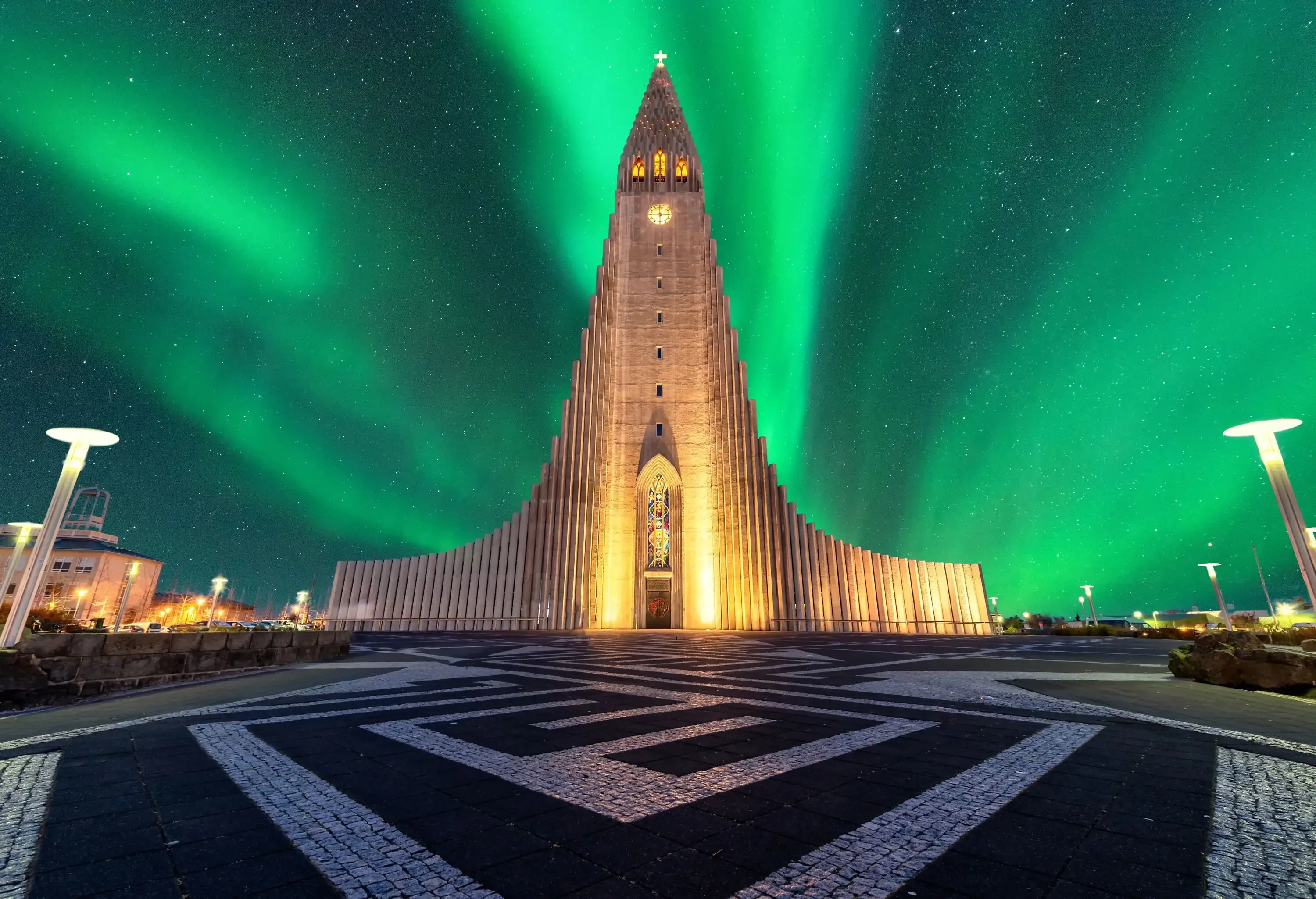A night-time view of Hallgrímskirkja, a church with curved spires and side wings under the green northern lights in a starry sky.