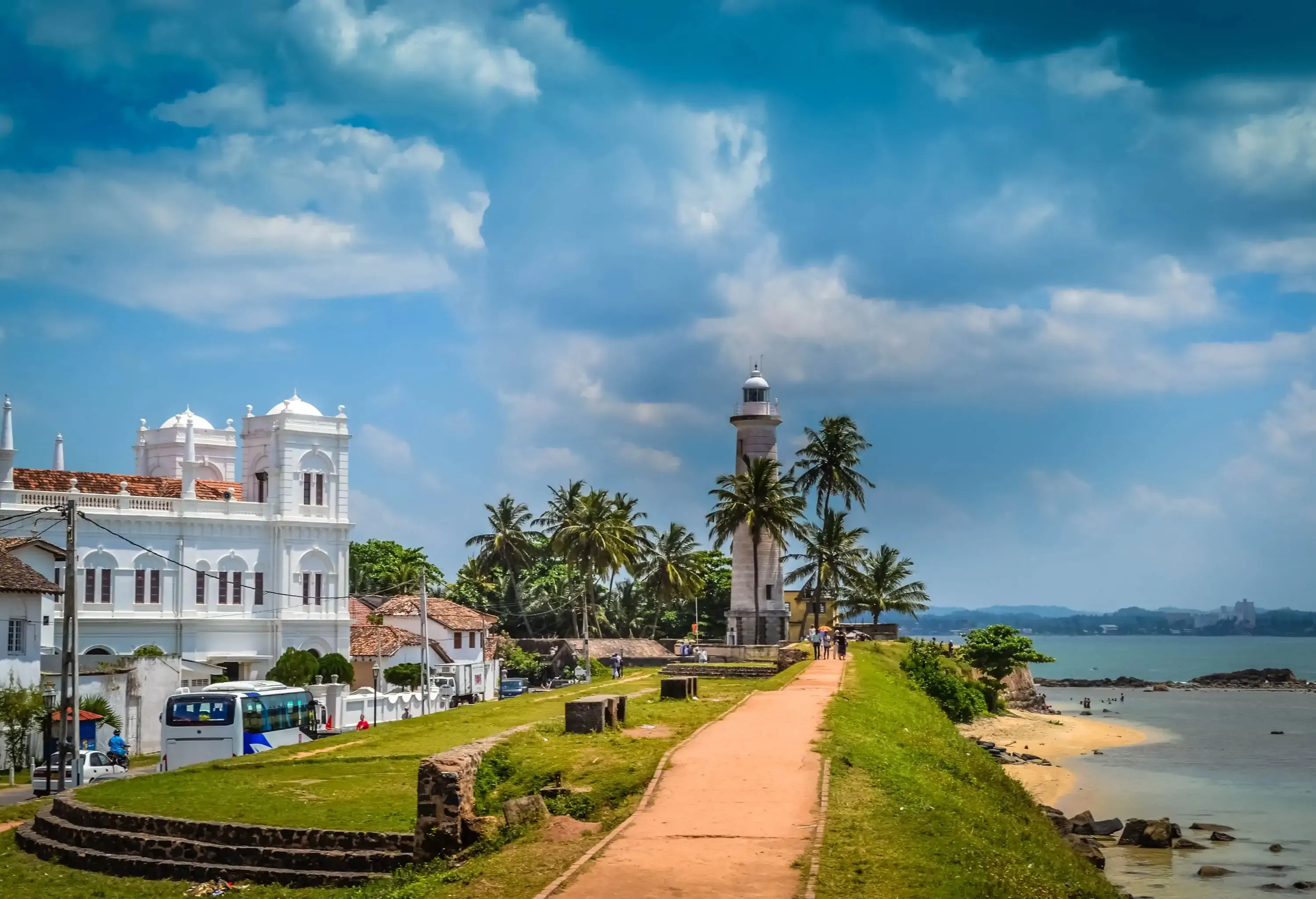 A paved walkway that traces the coastline and leads to a palm-covered lighthouse, with attractive houses dotting the sides.