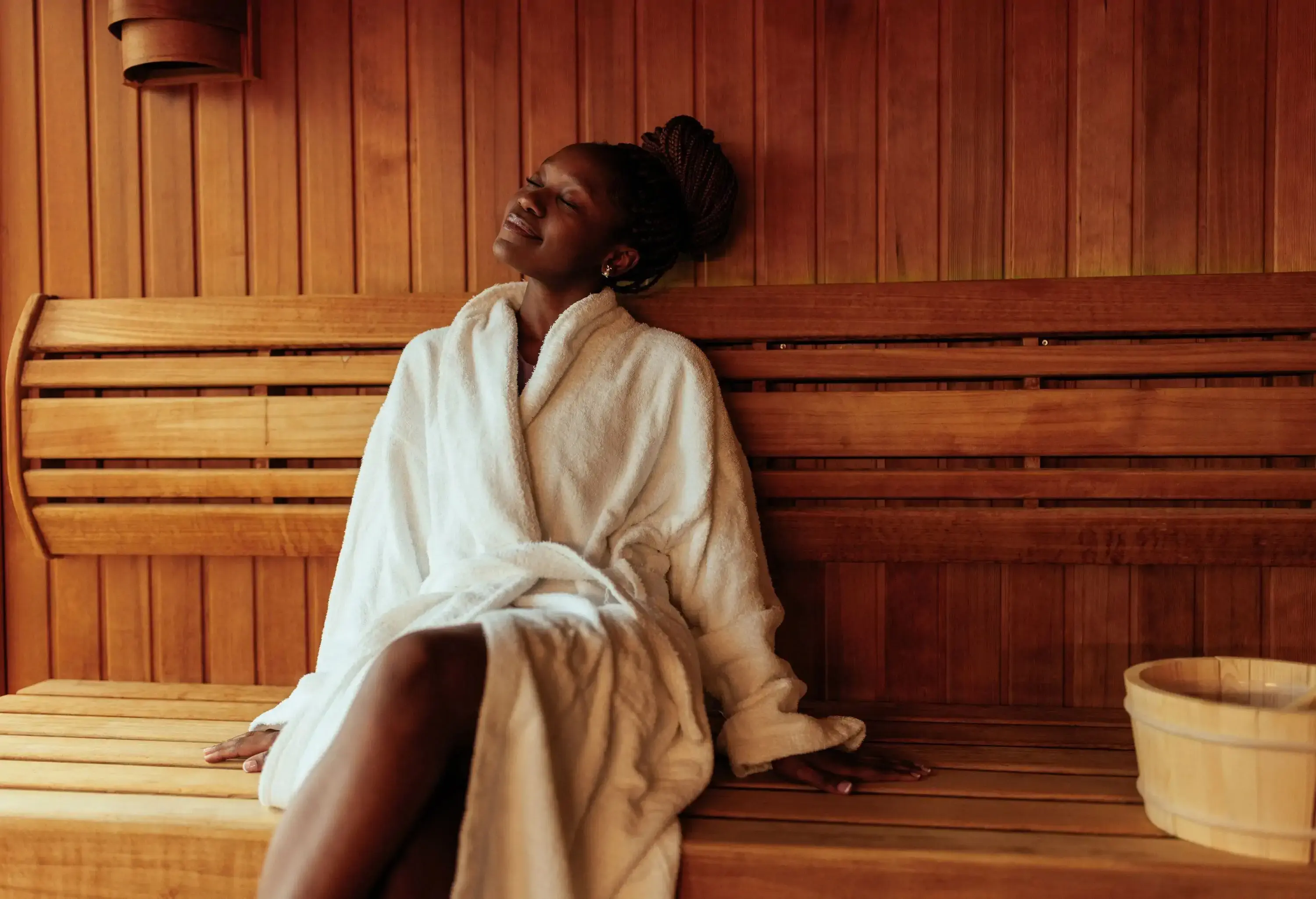 A young woman is sitting on the bench in the sauna wearing her bathrobe.