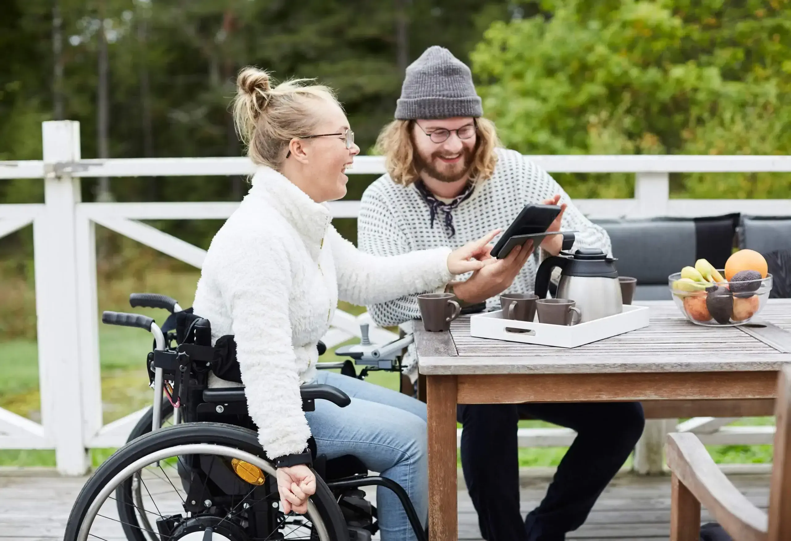 A happy woman in a knitted white long-sleeve sits in a wheelchair and points her finger at a mobile device shown to her by a man sitting next to her.