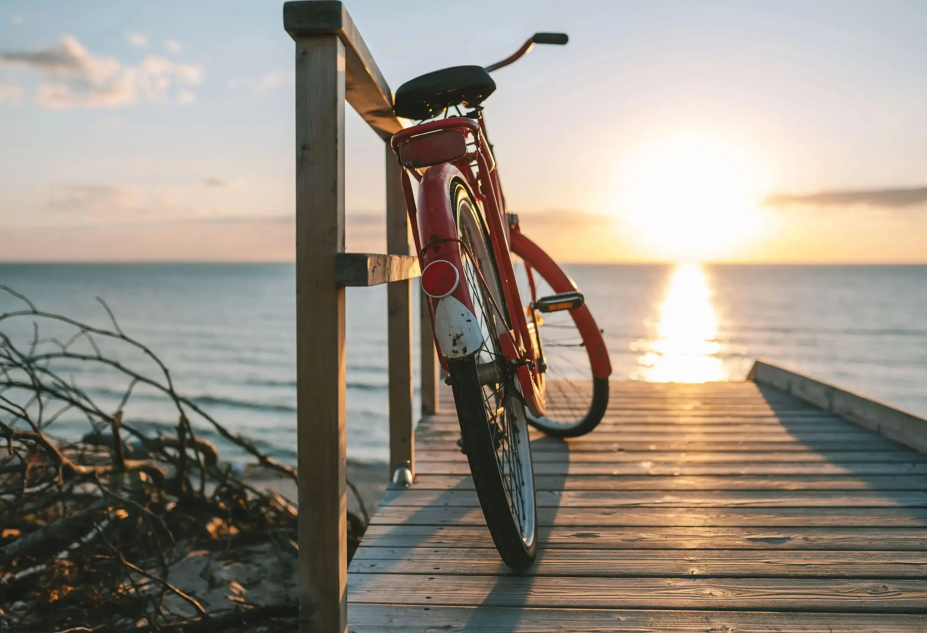 A red bicycle leaning on the wooden fence on a boardwalk alongside the sea with the sun shining brightly in the sky.