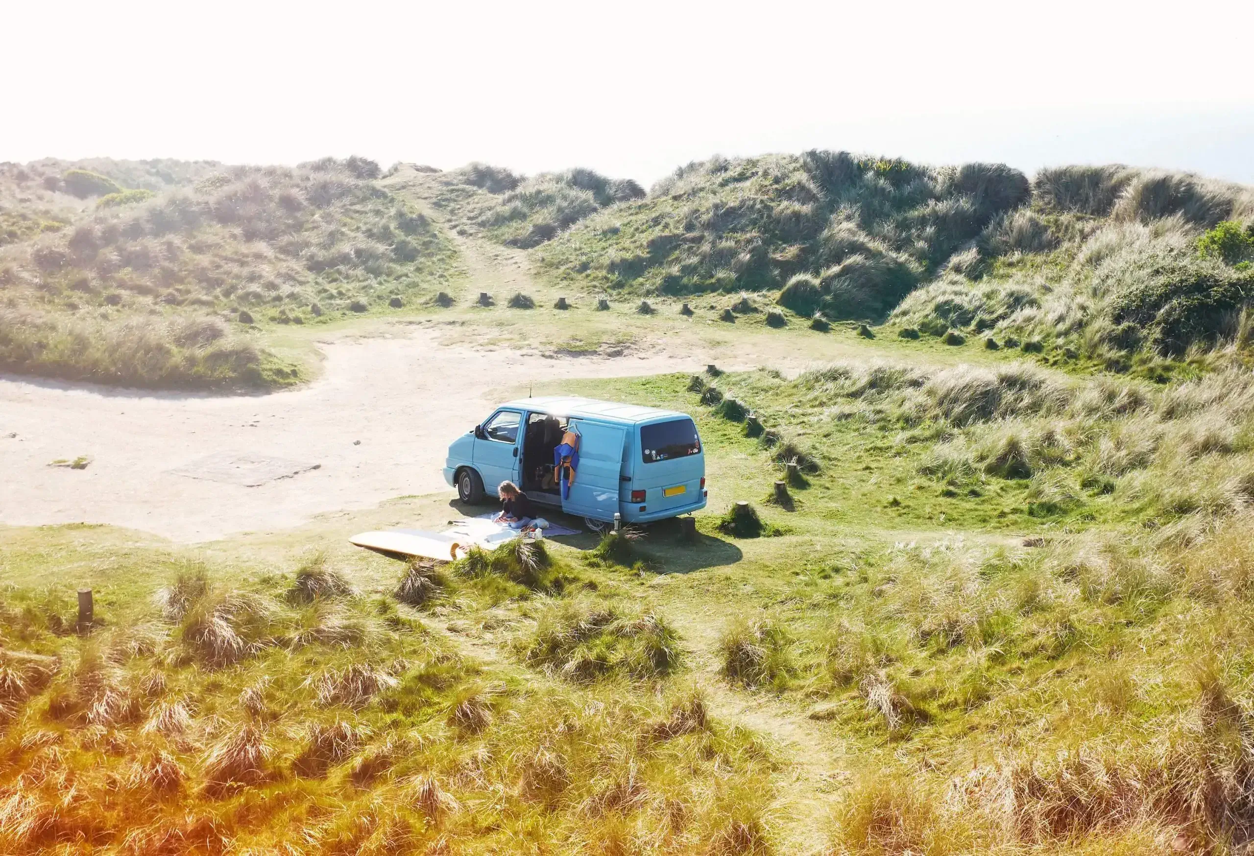 A person sits on a picnic mat beside a parked van in the grassy sand dunes.