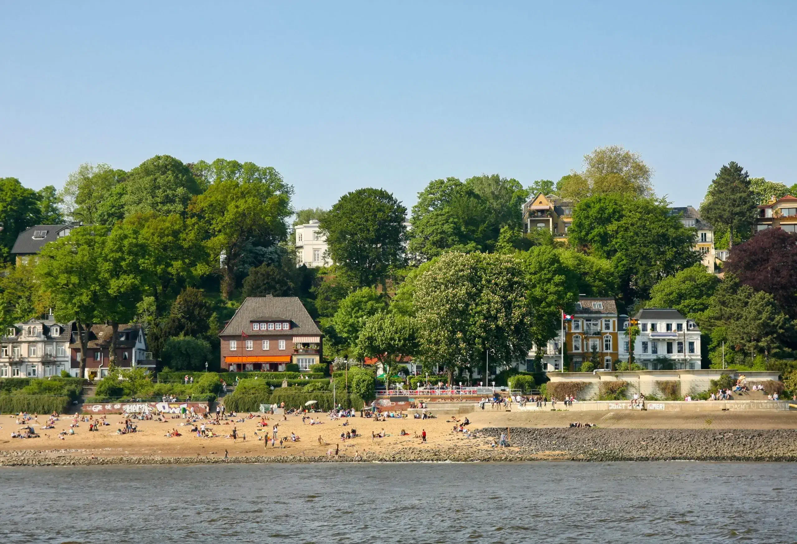 People wander on a brown sandy beach alongside houses surrounded by lush trees.