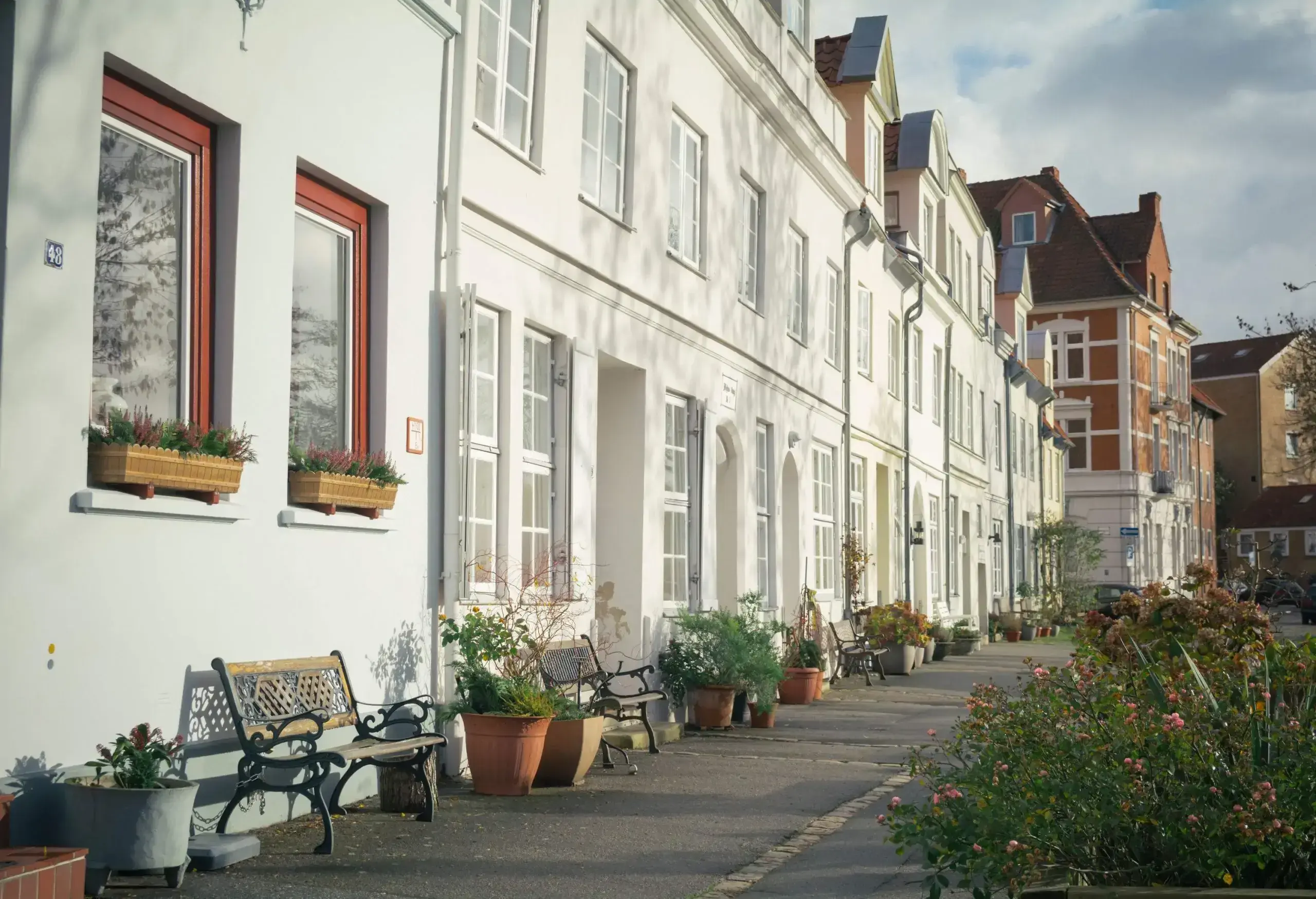 Bench chairs and potted plants outside of a white building along a small pathway.