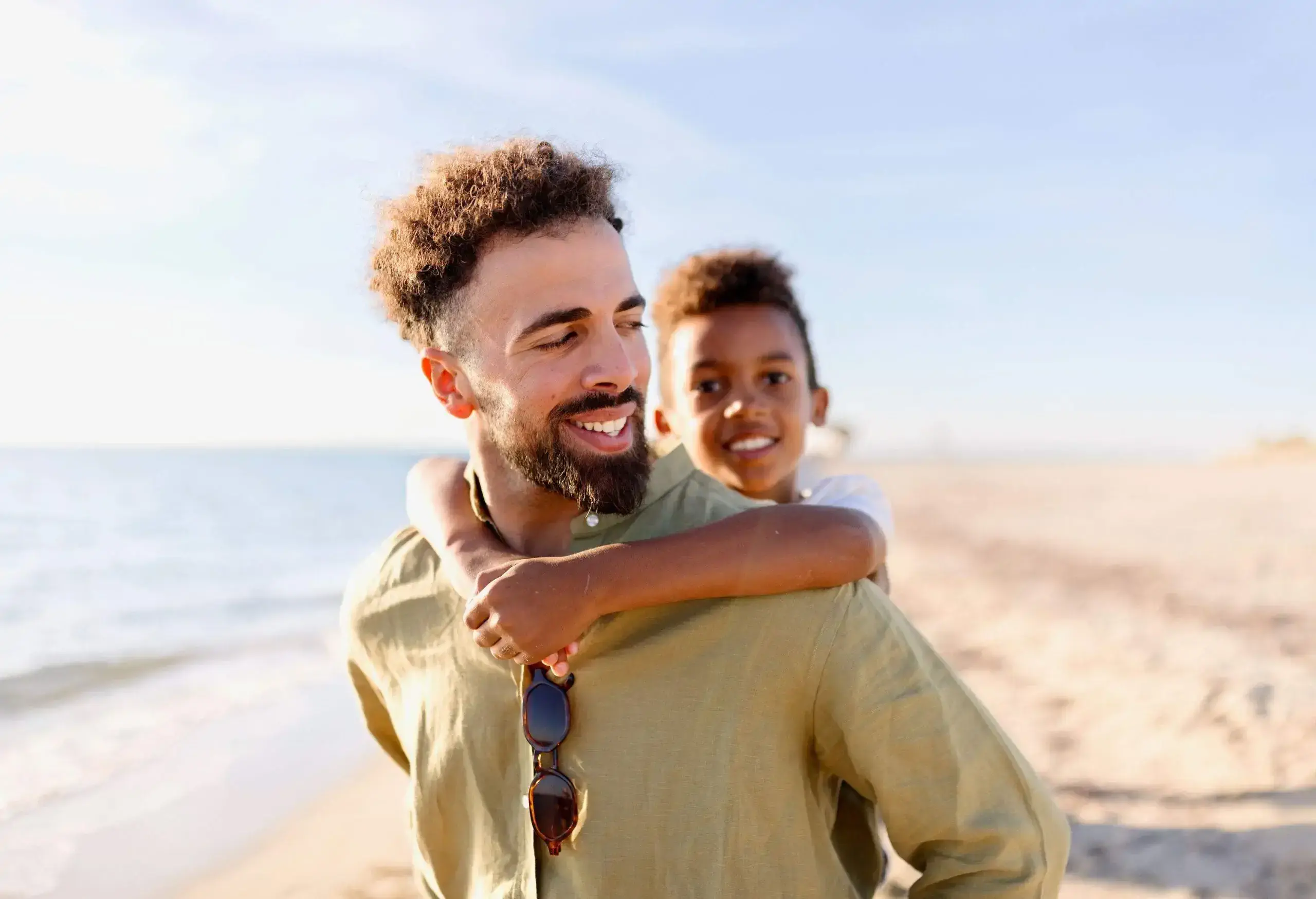 A man carrying a little boy on his back while walking on the beach.