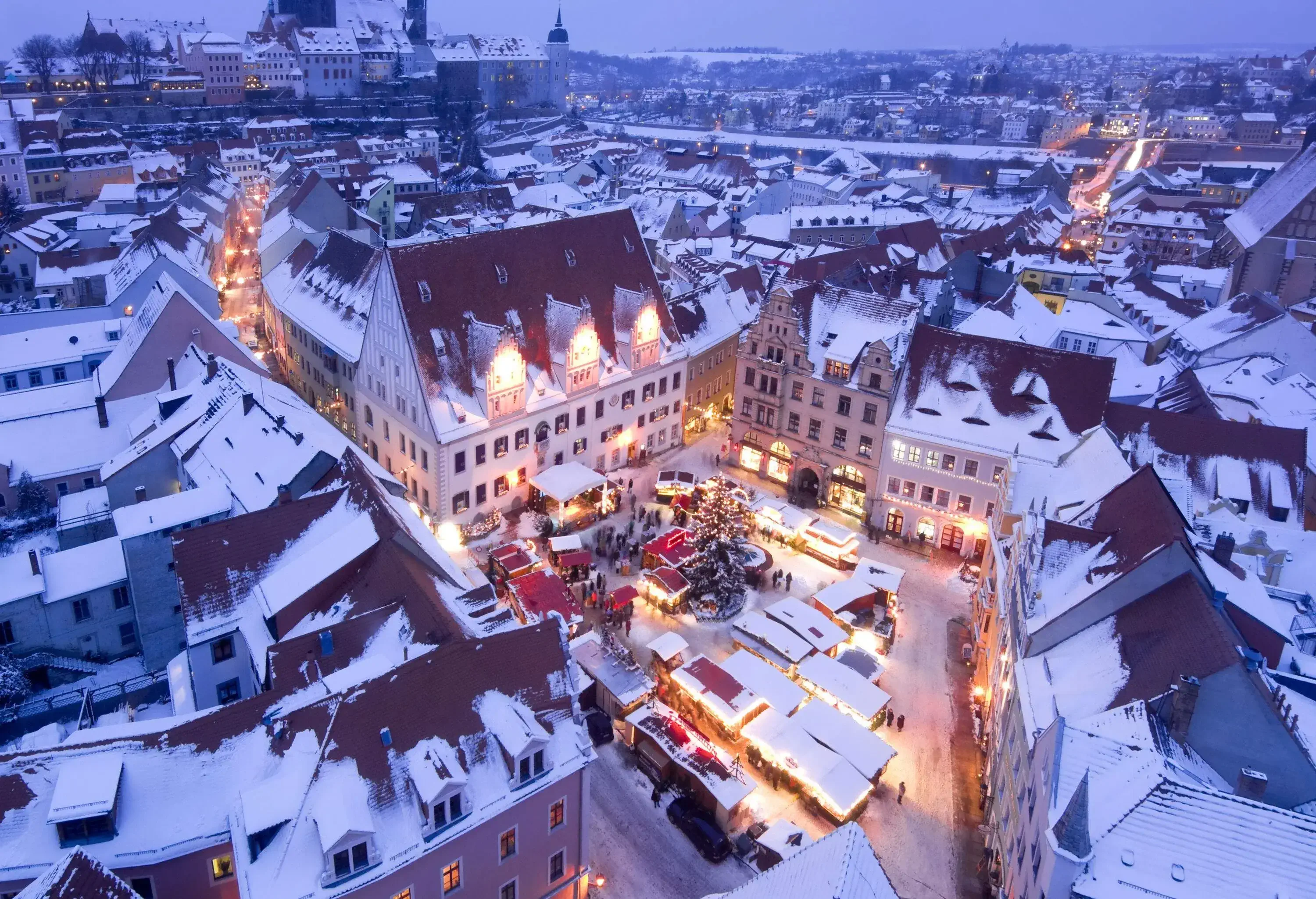 A town plaza covered in snow with Christmas shops decorated with lights.