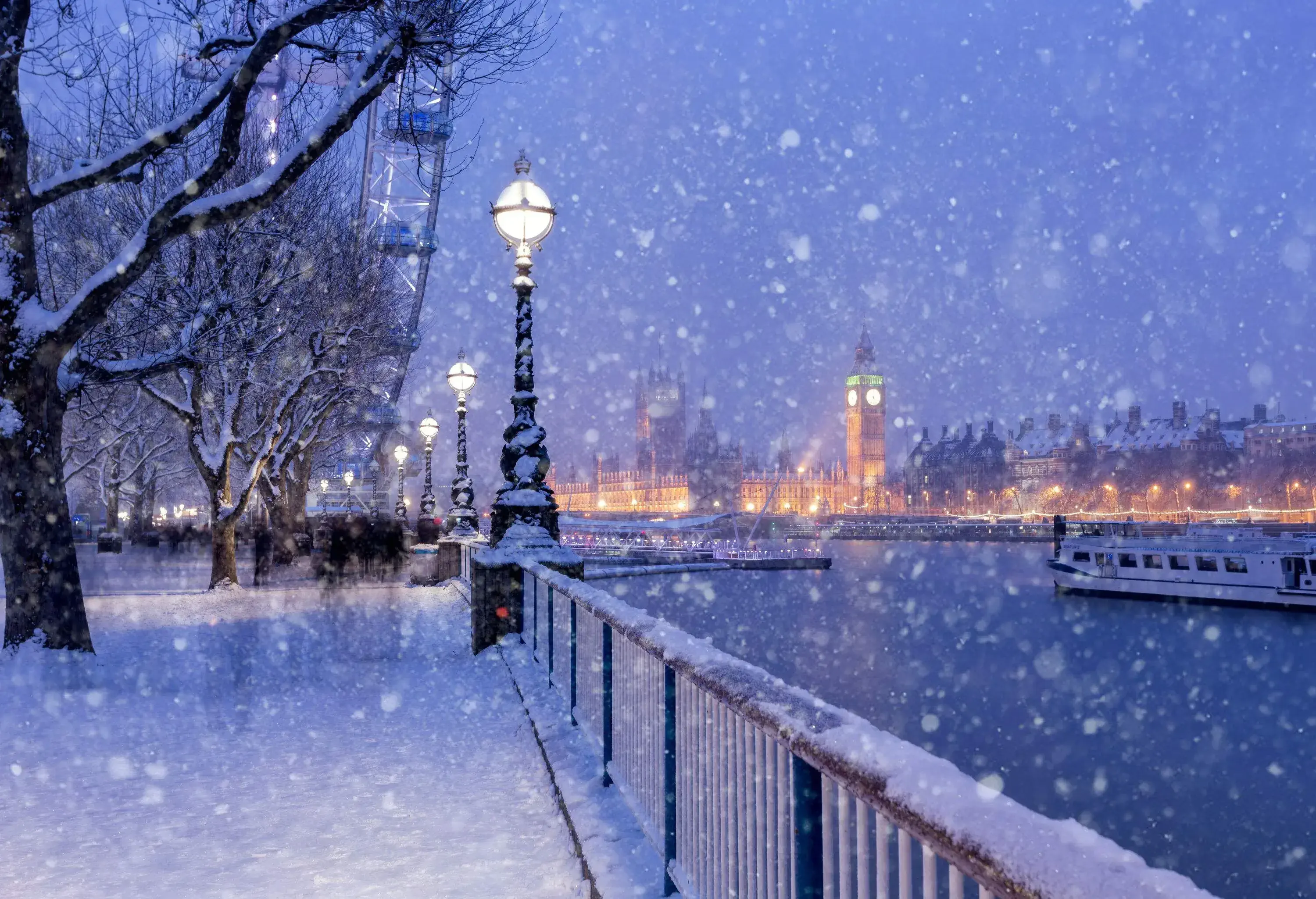 A snow-covered promenade by the river lined with tall bare trees and illuminated lamp posts on a snowy winter night.