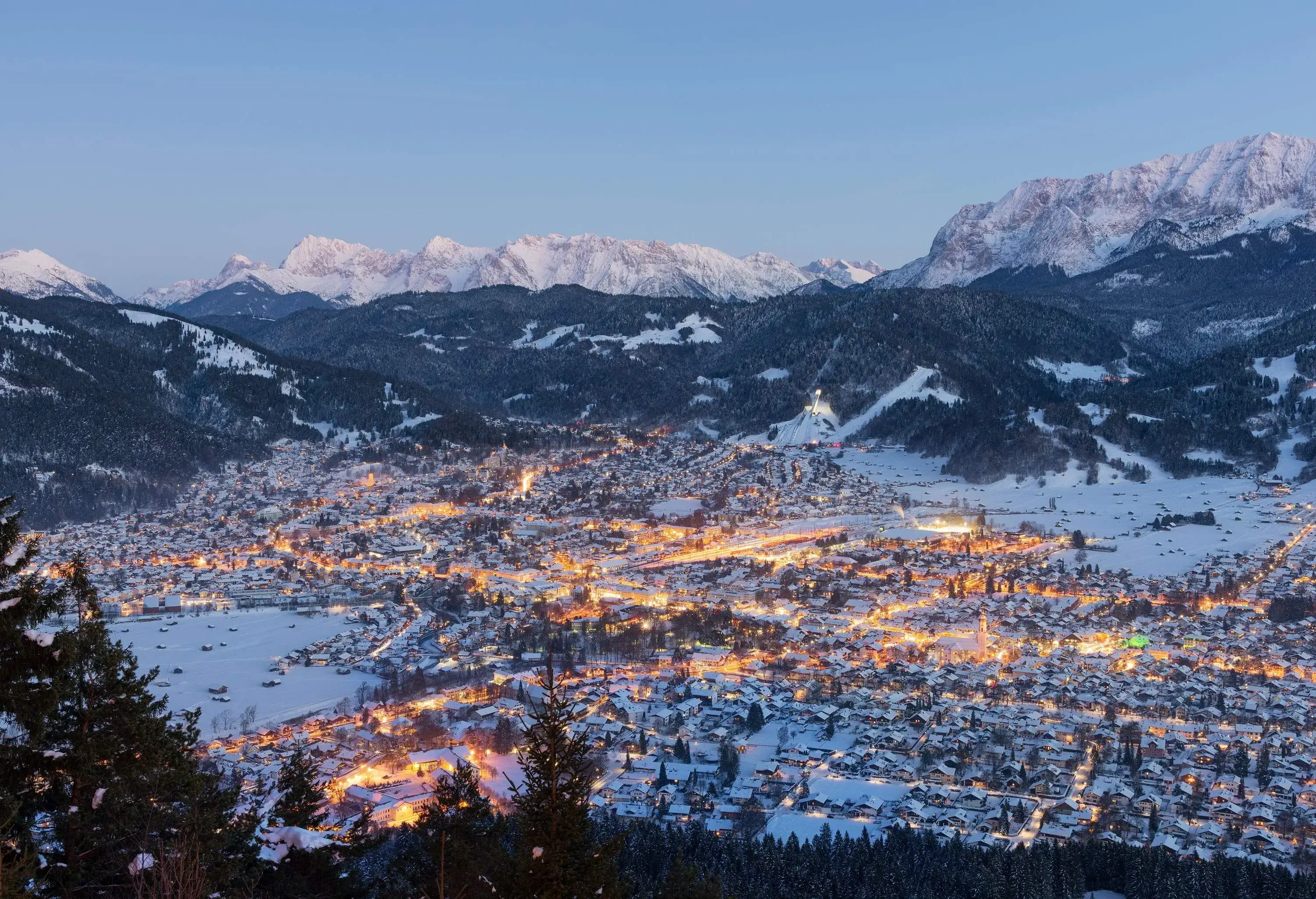 Scenery of a snow-covered forest surrounding an illuminated town at night.