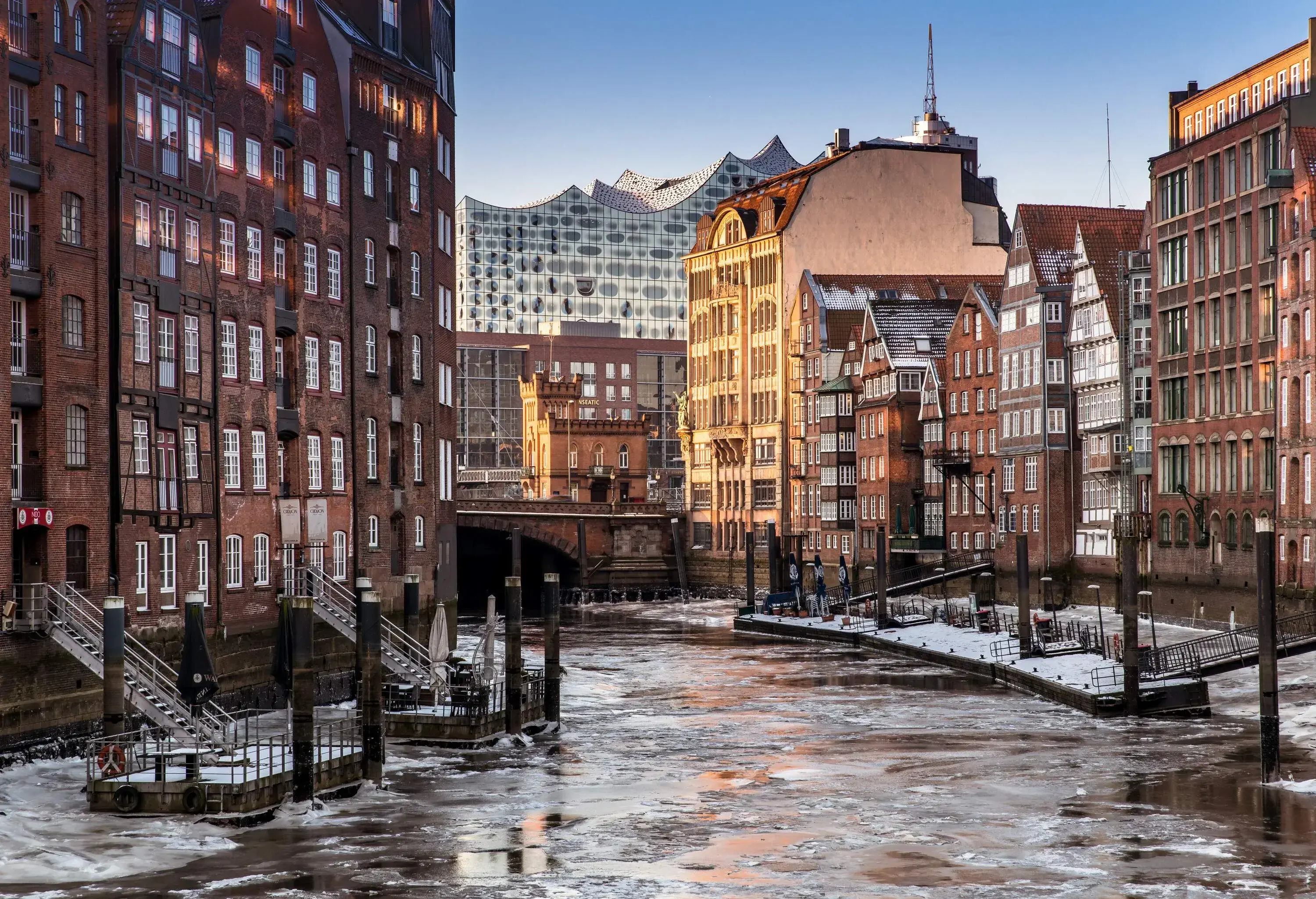 A frozen canal passes through a brick arch bridge alongside docks in the middle of tall warehouse buildings.