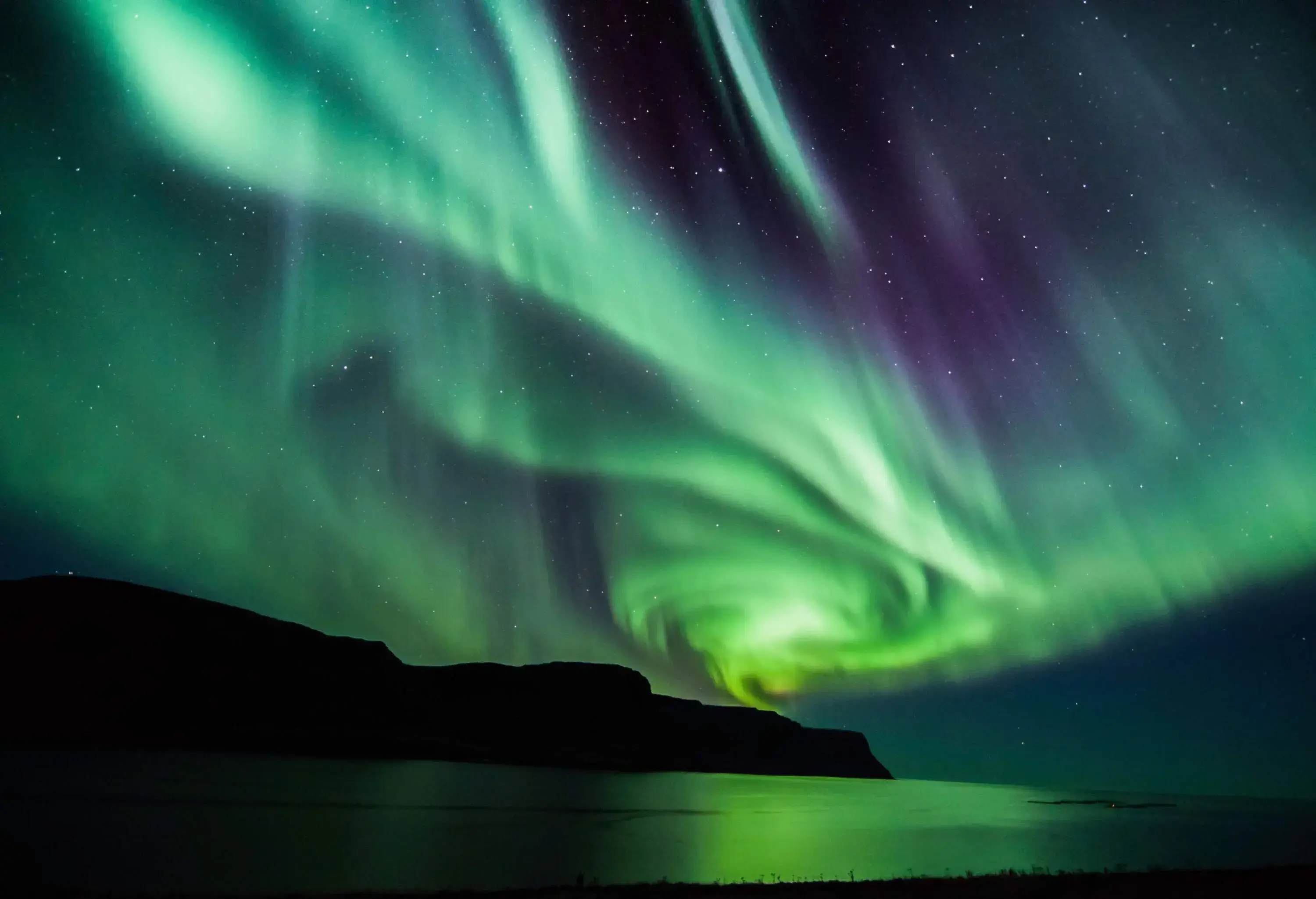 Mesmerising northern lights shine above the sea, surrounded by cliffs on a dark night.