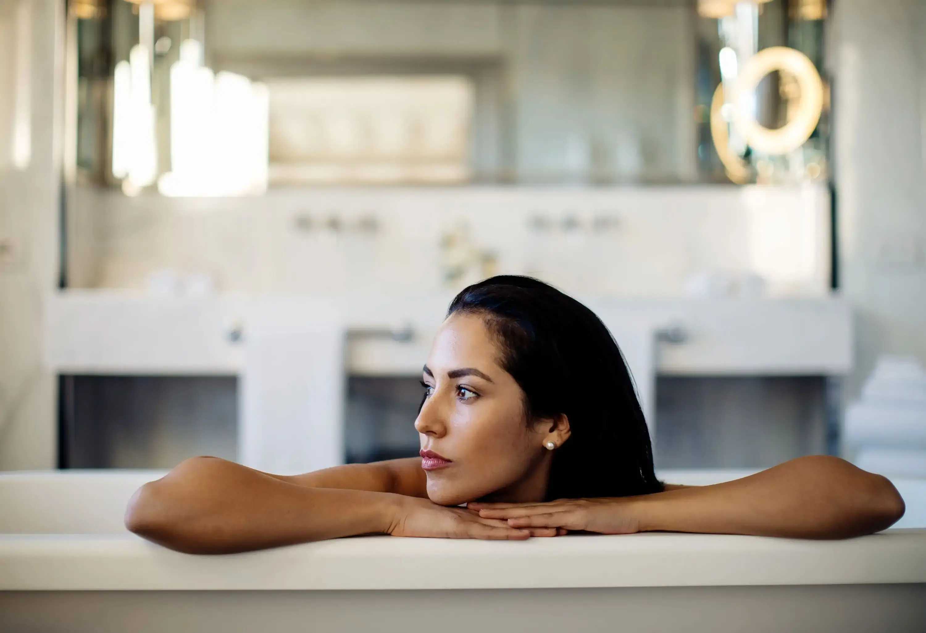 A woman submerged in a bathtub inside a brightly lit bathroom, looking to her right side.