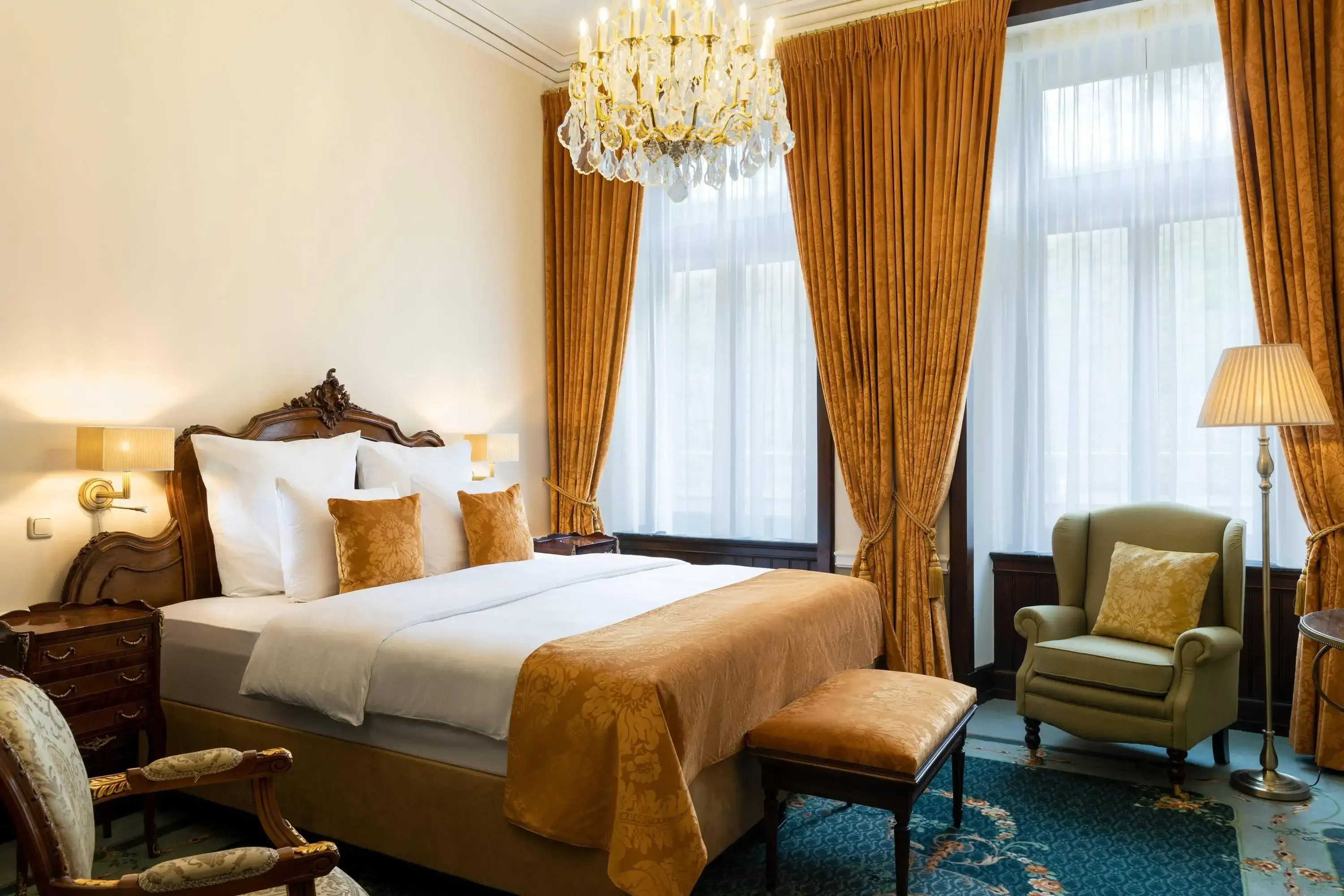 A luxurious hotel bedroom with a grand wooden bed, gold accents, tall drapes, and a crystal chandelier.
