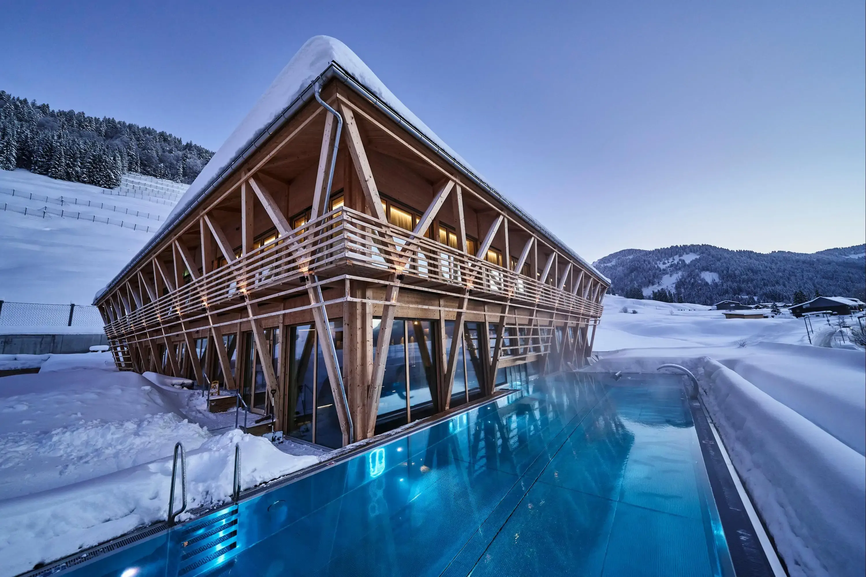 A wooden house surrounded by snow, featuring a swimming pool in the foreground, creating a serene winter scene.