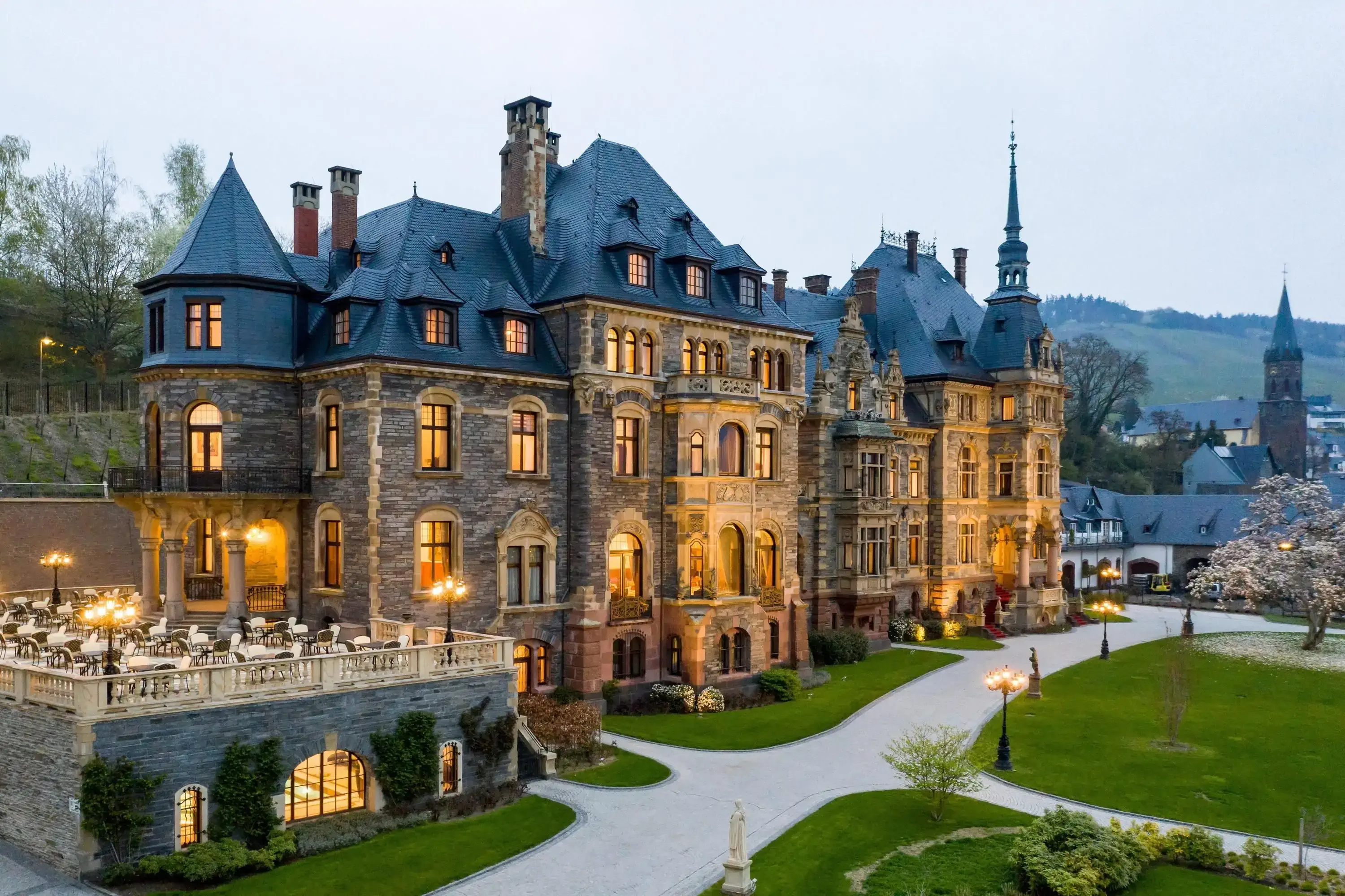 A grand chateau with a mix of stone and ornate architectural details. The building features towers, balconies, and arched windows, surrounded by  lawns and a terrace with dining tables. 