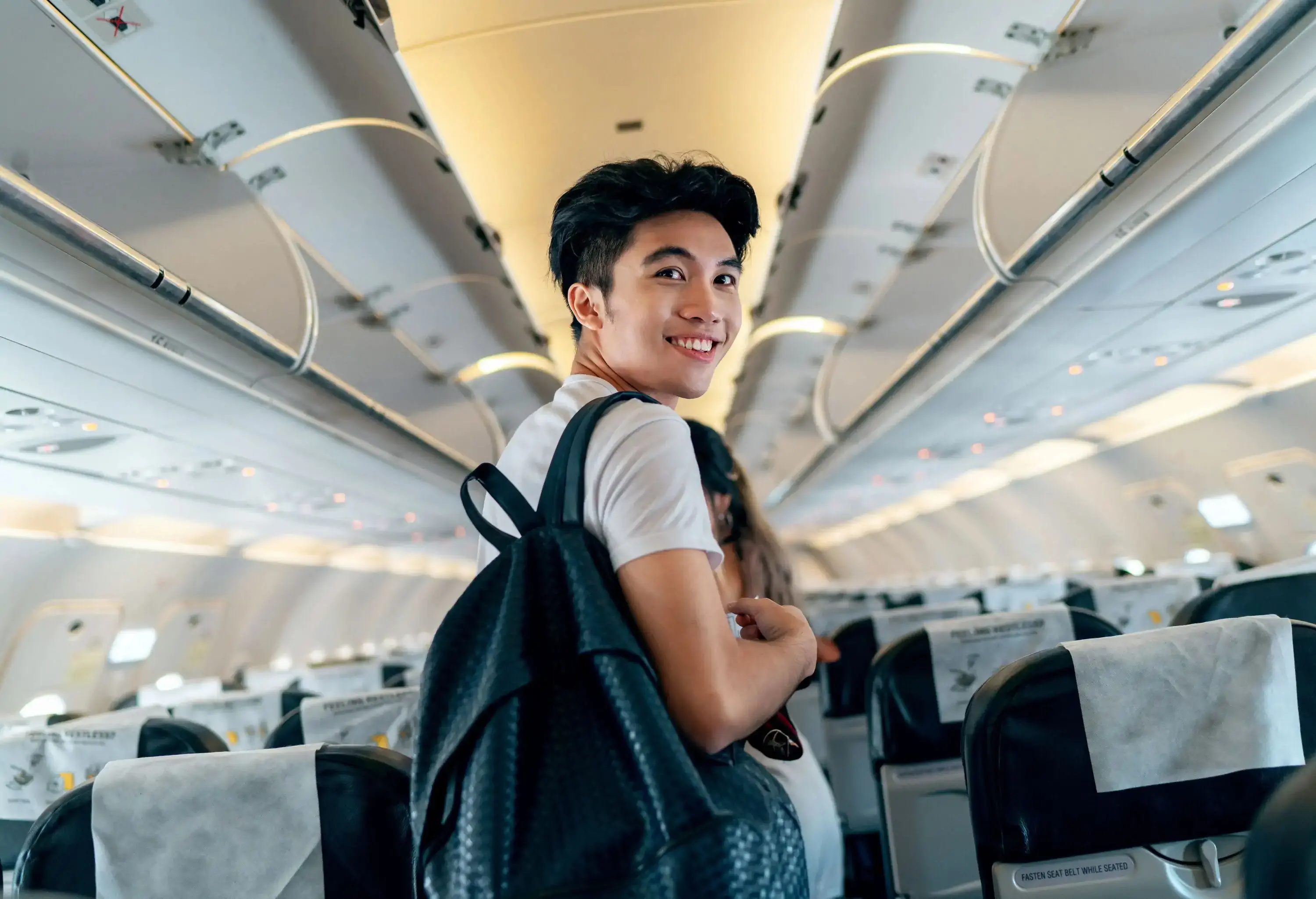 A young male traveller boards a plane, carrying a backpack and glancing back.