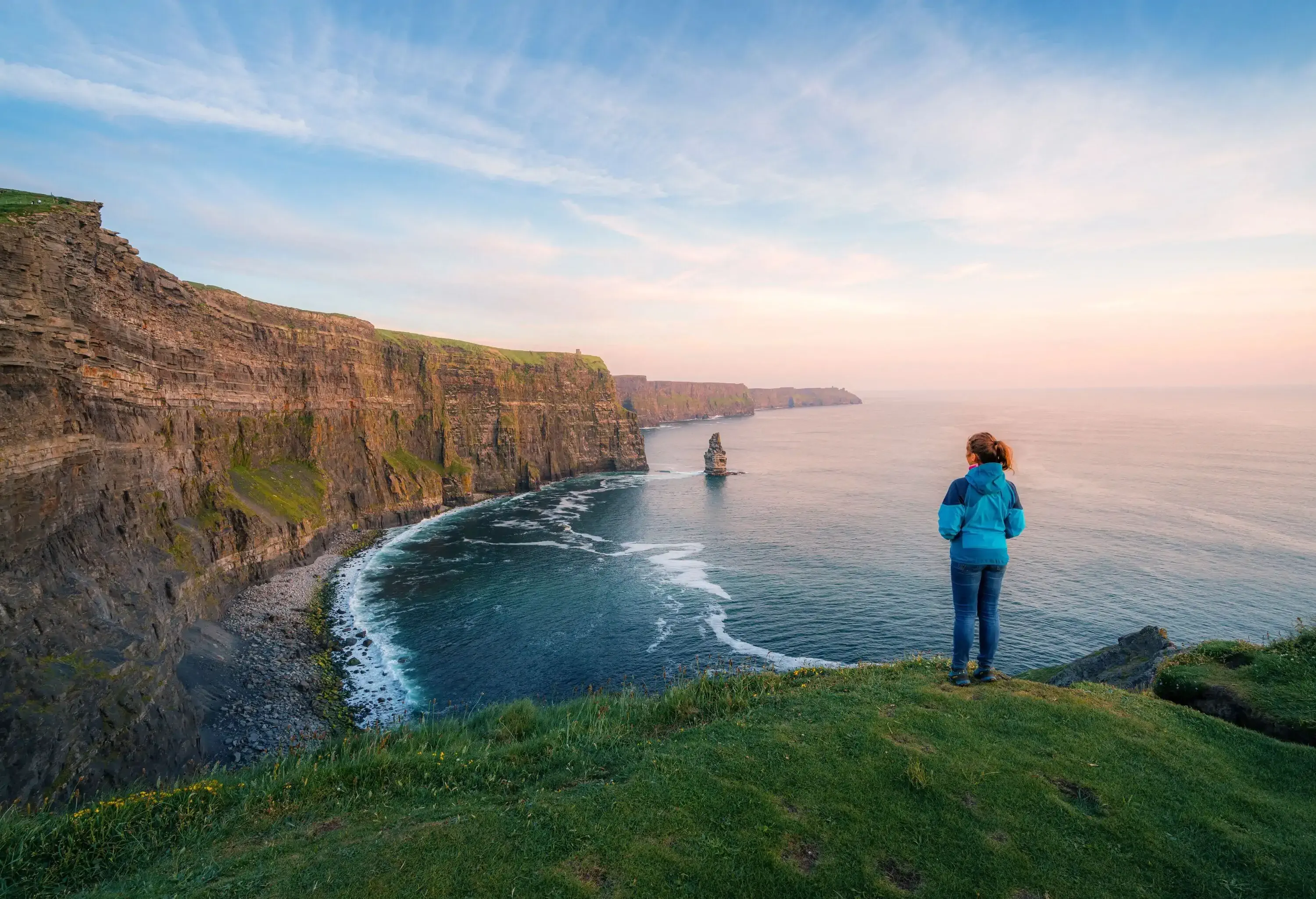 A woman in a blue jacket gazing across to the entrancing Cliffs of Moher while perched on a ledge.