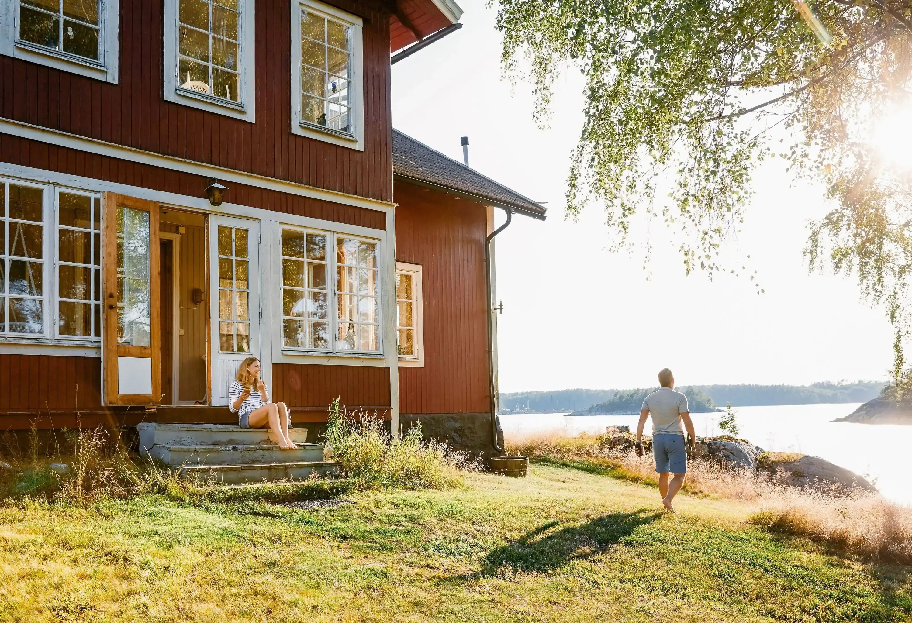 Man walking by typical red Swedish lake house whilst a woman is sitting on the stoop