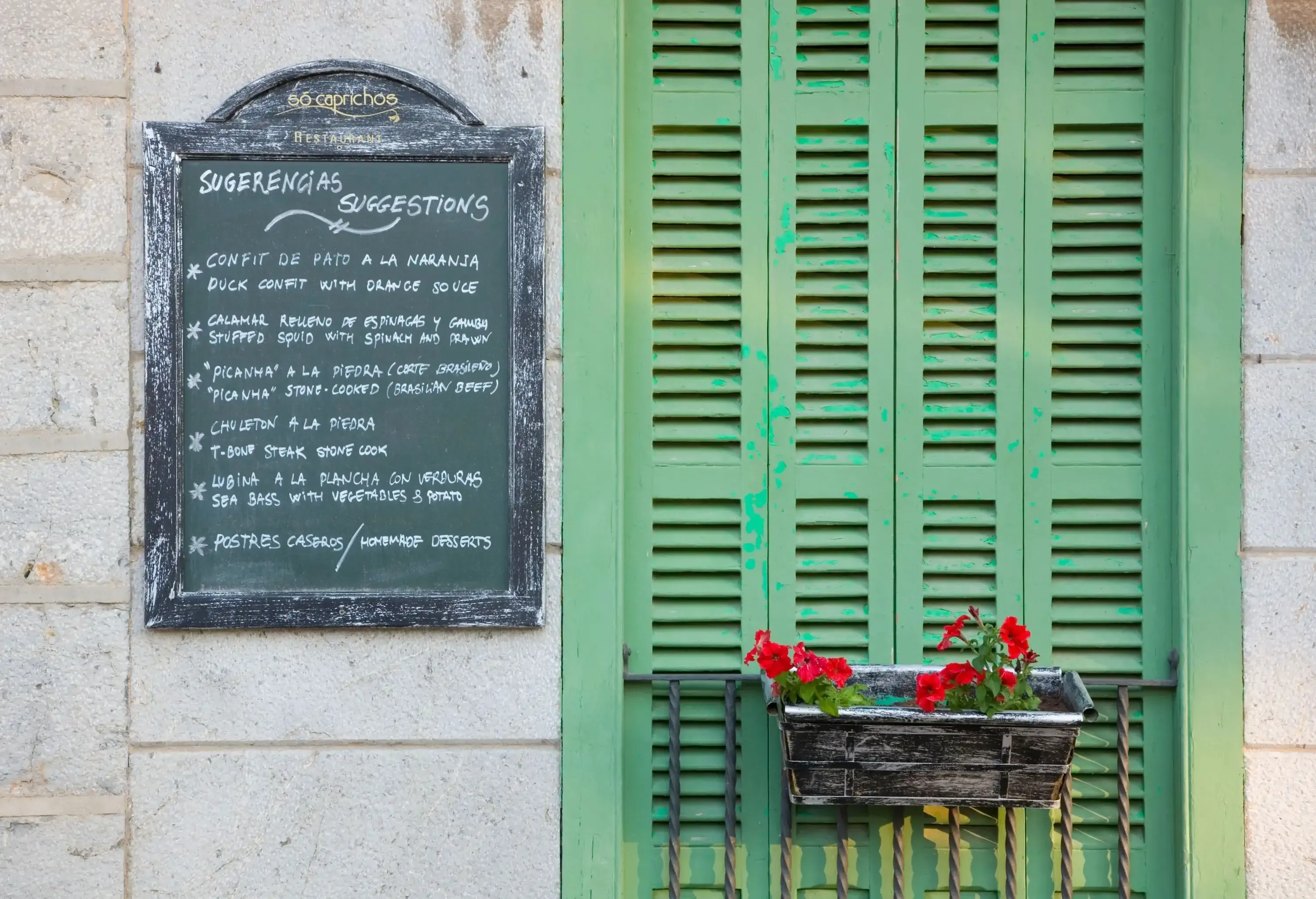 A restaurant menu written on a blackboard hanging next to a green shutter of an old building with red geraniums in a vase