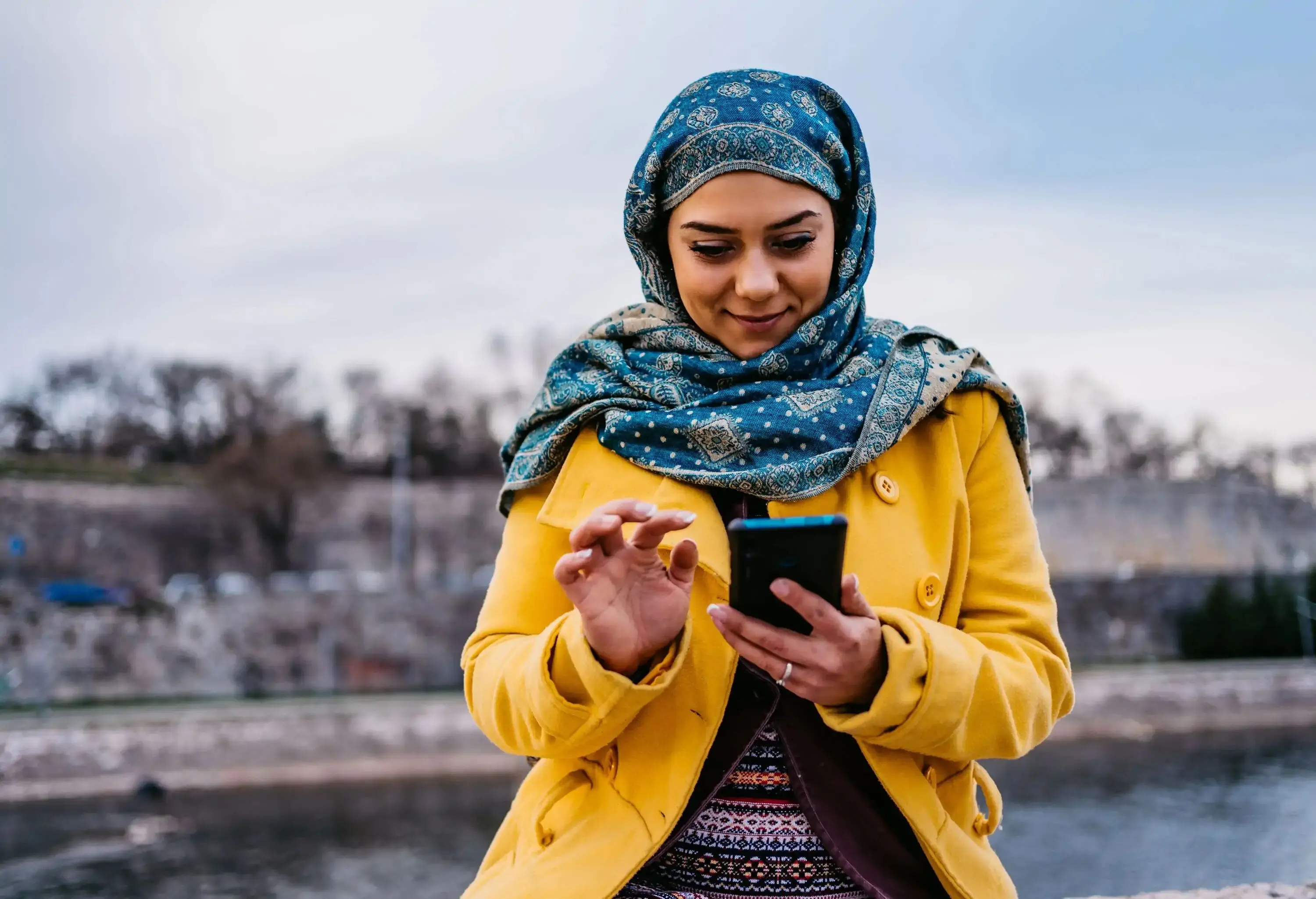A person wearing a headscarf and a yellow coat smiles while using their smartphone.