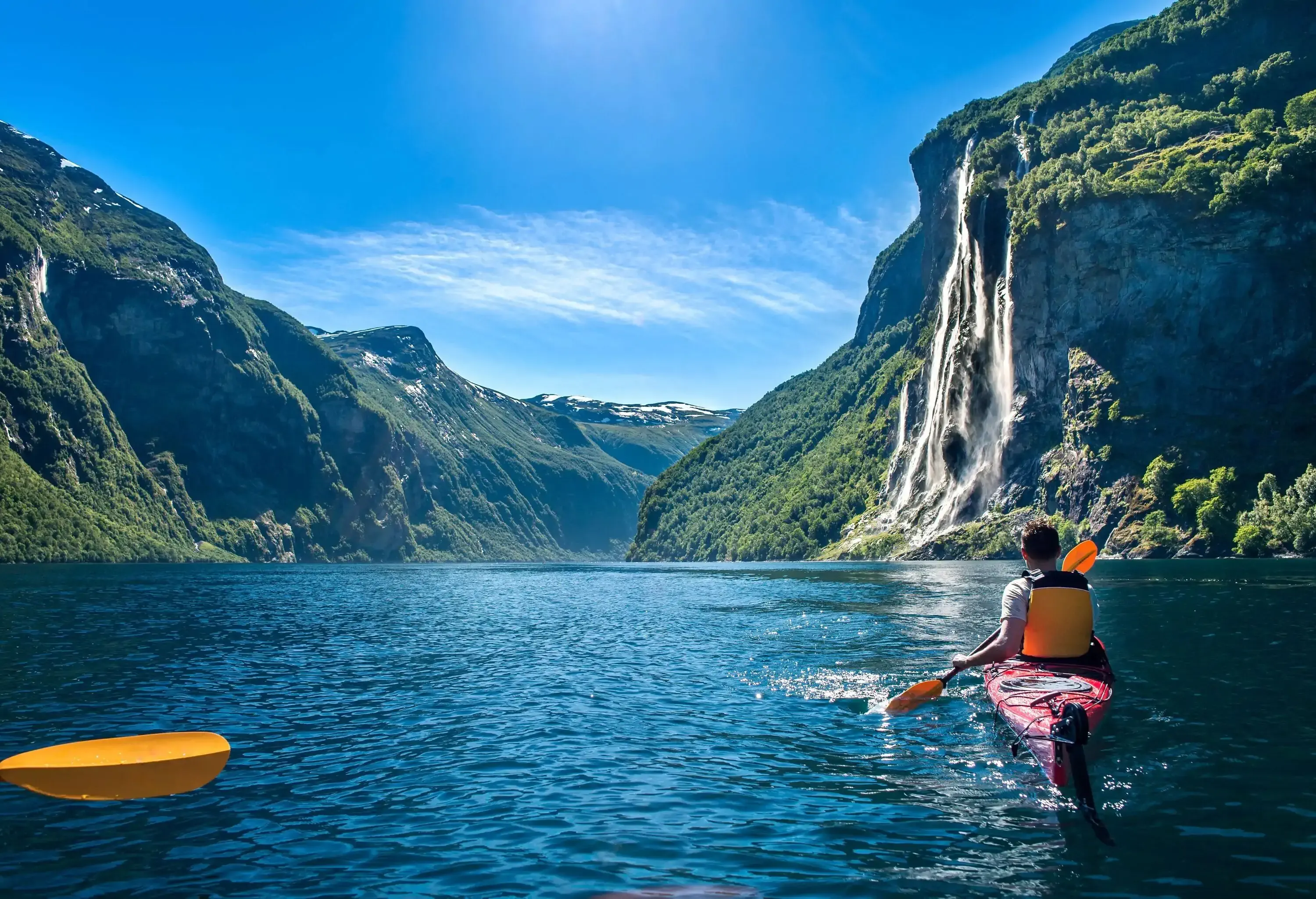 A man in a kayak paddling towards a waterfall on the mountainside on a tranquil lake surrounded by high mountains.