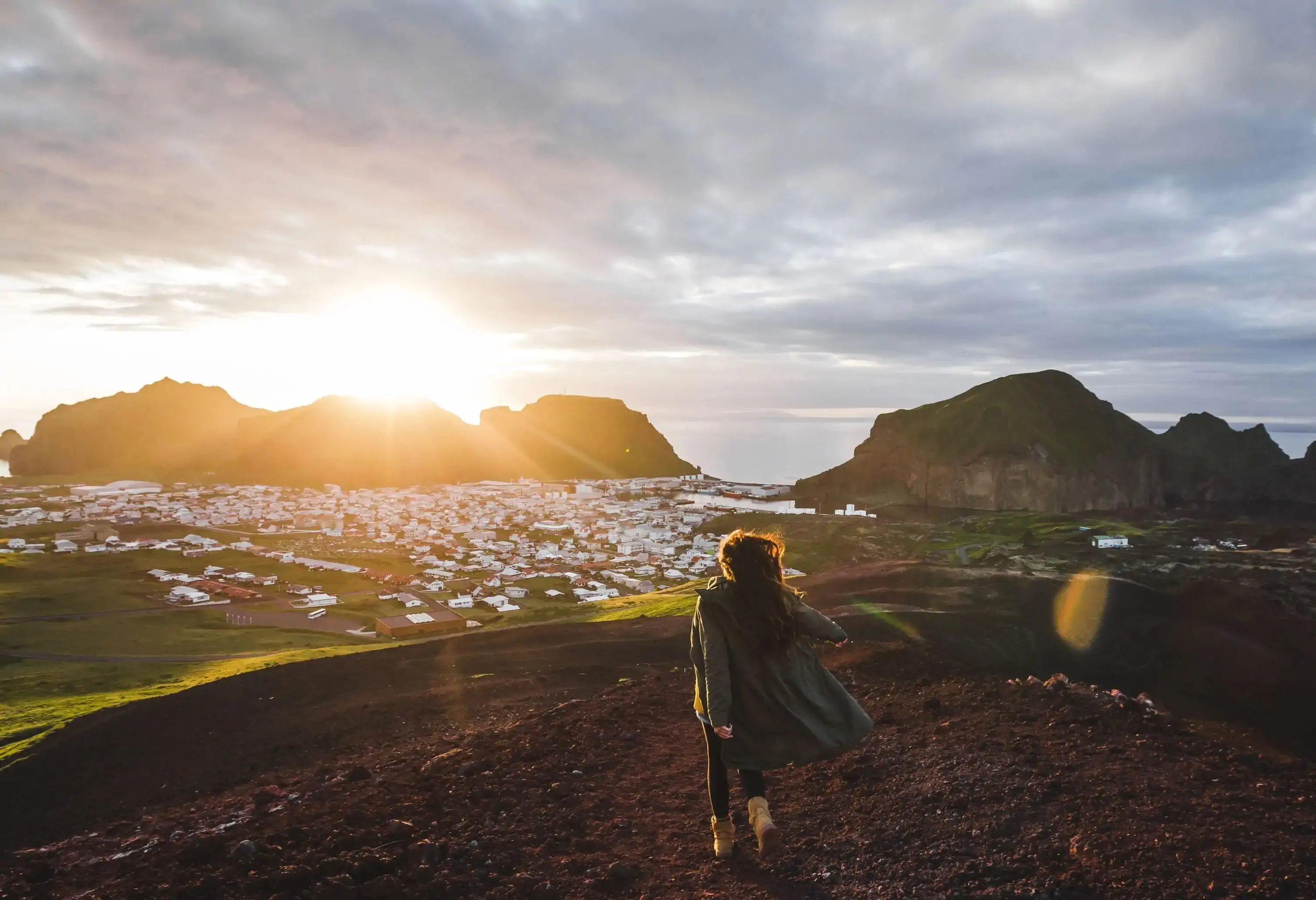 A young woman in a long coat stands on the summit of Eldfell volcano, bathed in the glory of a beautiful sunset, with a breathtaking panoramic view of the white-washed settlement below.